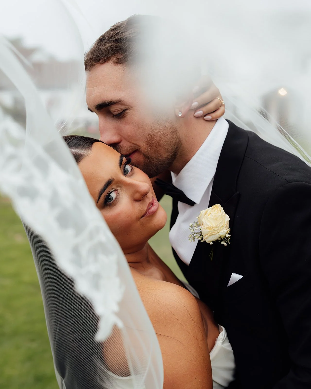 Close-up portrait of Jack kissing Nisha’s forehead beneath her flowing veil at Batch Country House in Somerset, UK, with Nisha looking towards the camera and Jack’s white rose buttonhole visible, photographed naturally by Just James Weddings.