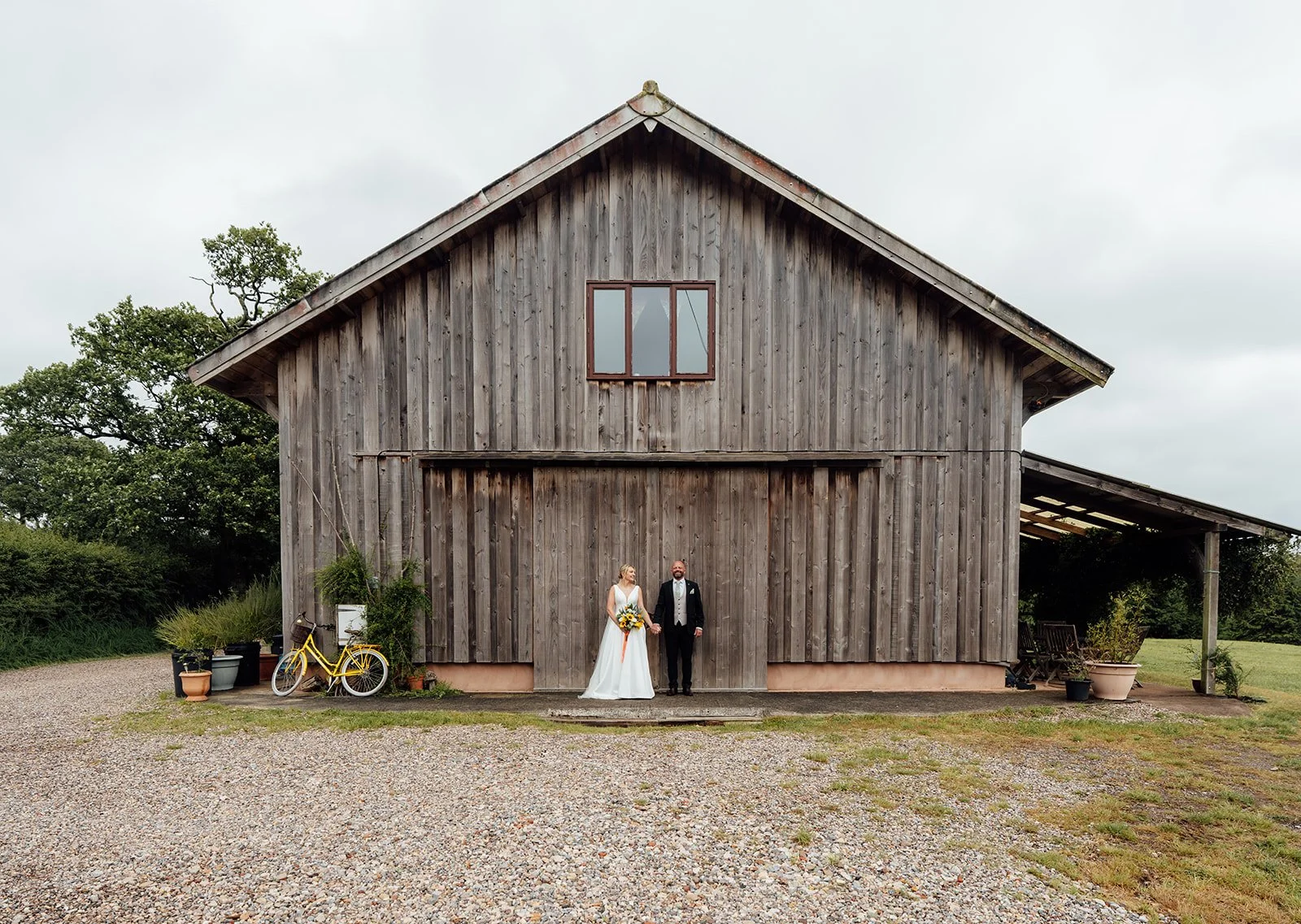 Wide wedding portrait of Hannah & Ben at Grays Barn in Devon, UK, standing hand in hand in front of the rustic timber barn, with a yellow bicycle and countryside details adding relaxed character, photographed by Just James Weddings outdoors.