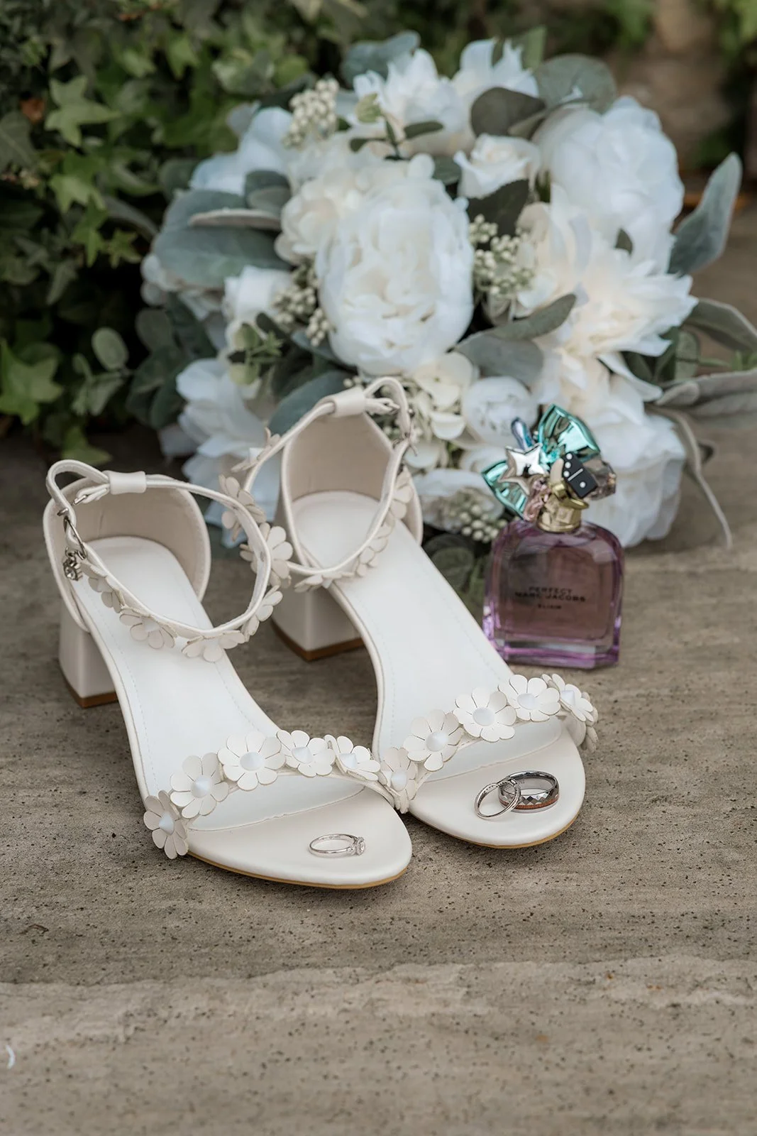 White bridal shoes with floral straps, wedding rings, perfume bottle and white bouquet styled on stone steps at Siobhan & Nicholas' wedding at Kingscote Barn, Cotswolds, UK, elegant wedding details from a romantic rustic barn wedding morning.