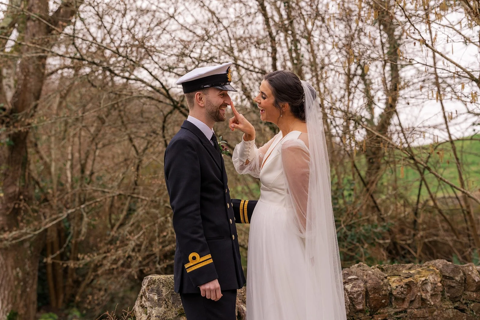 Megan playfully taps Luca on the nose as they laugh together beside an old stone wall at The Great Barn in Devon, Luca dressed in his Royal Navy uniform with gold-braided epaulettes and Megan in her elegant long-sleeved wedding gown and veil.
