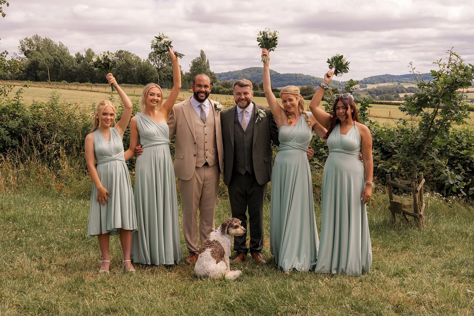Aaron & Jack stand with their bridesmaids and dog Milo as bouquets are raised during group photos at Rock Village Hall in Worcestershire, photographed by Just James Weddings.