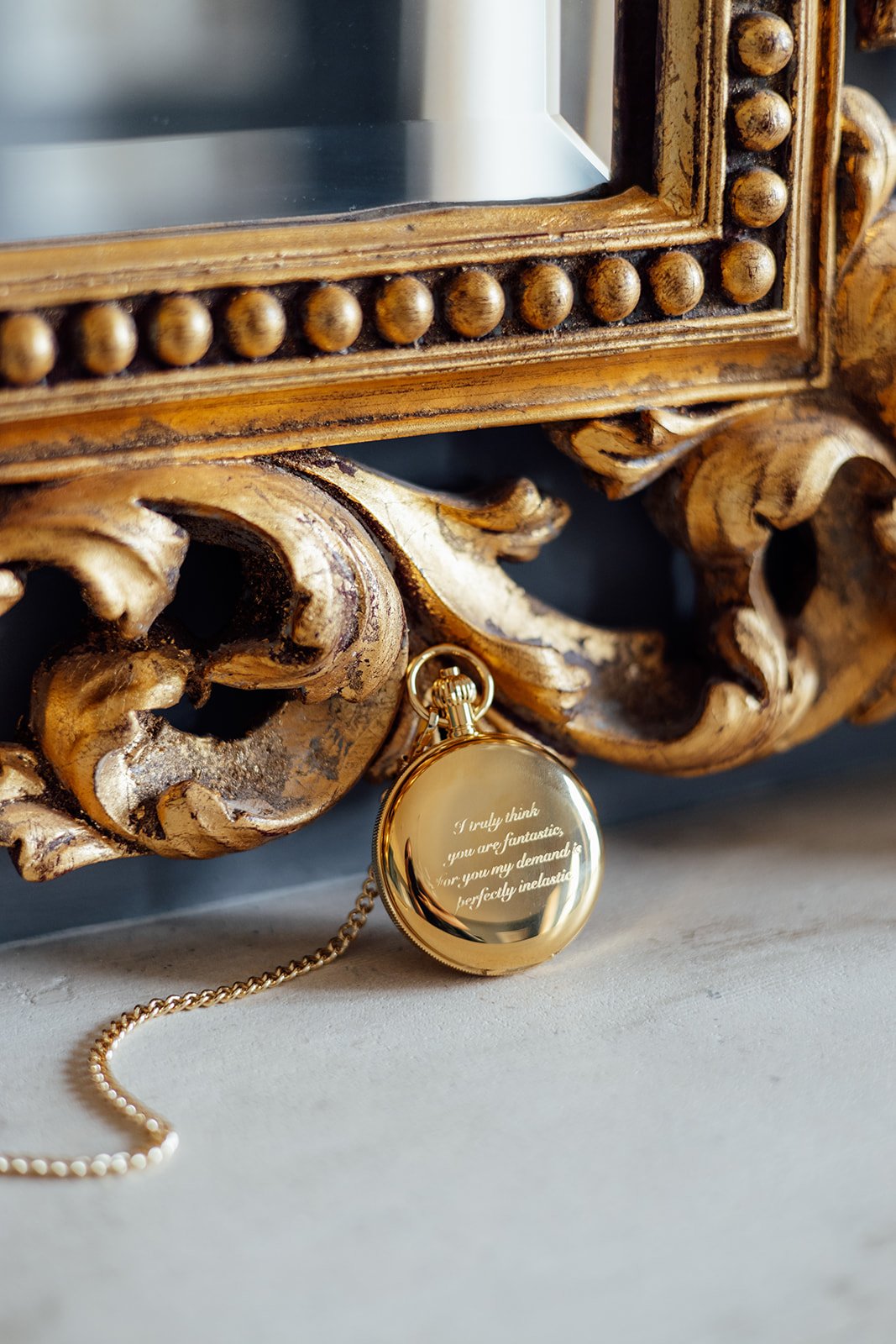 Gold pocket watch displayed beside an ornate gilded mirror at Caitlin and Kevin’s Clevedon Hall wedding in Somerset, featuring a personalised engraved message on the cover.