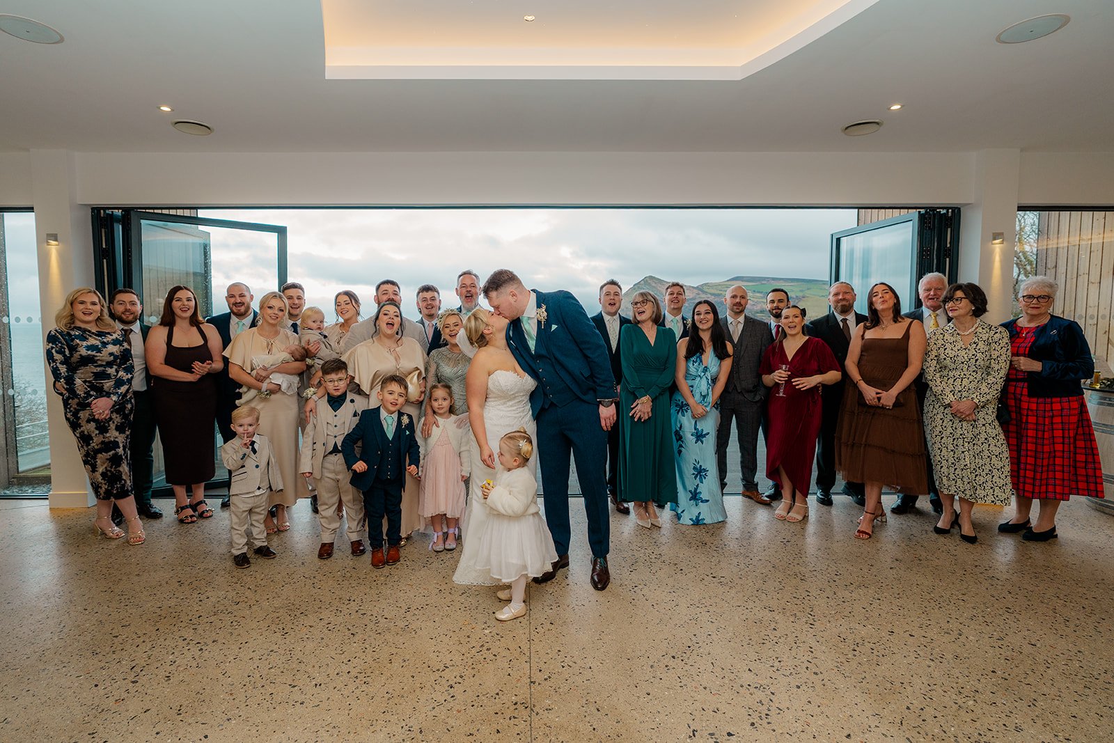 Andrew & Daisy sharing a kiss surrounded by family and friends at Sandy Cove Hotel in Devon, framed by panoramic sea views through open doors, capturing a relaxed group wedding portrait at this modern coastal venue.