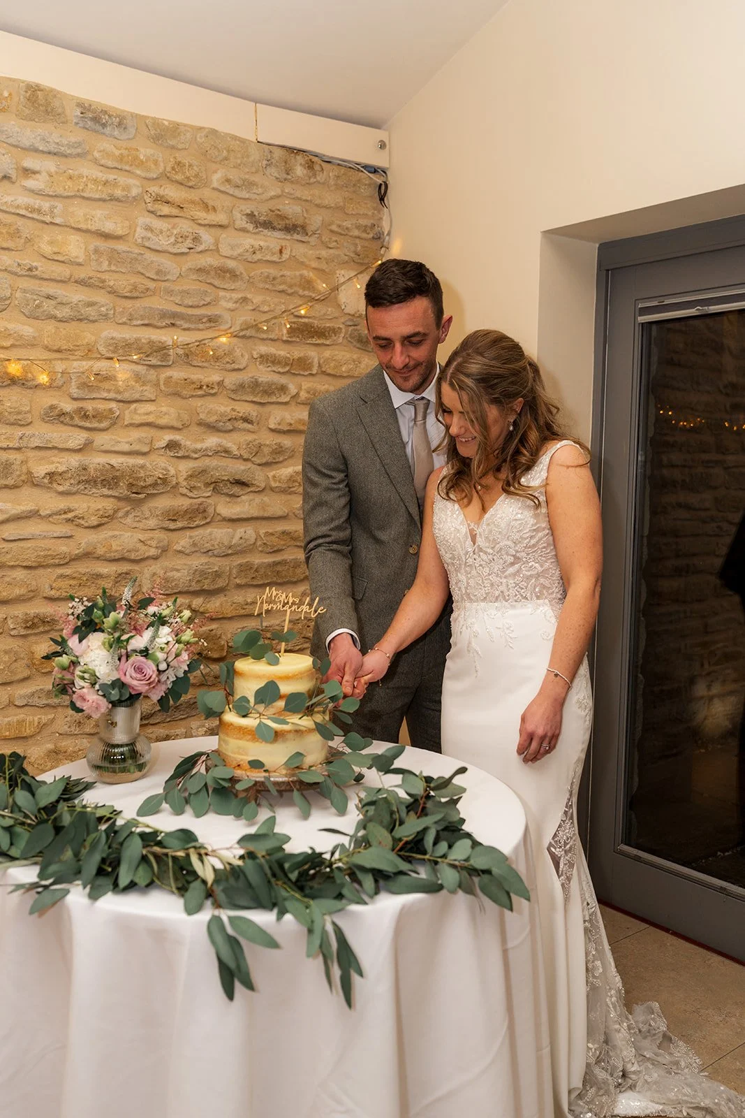 Anna and Tom cut their wedding cake at Winkworth Farm in the Cotswolds, UK, beside soft florals and greenery. This indoor reception photo captures the couple smiling together by a semi naked cake in warm, rustic Cotswolds wedding photography.