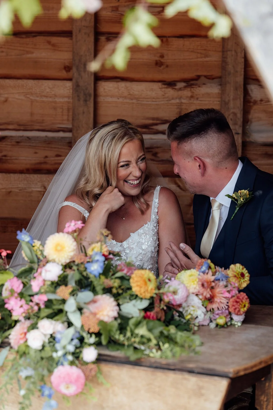Elise and Tom laughing together behind colourful wedding flowers during their outdoor ceremony at Bowerchalke Barn in Wiltshire, photographed by Just James Weddings.
