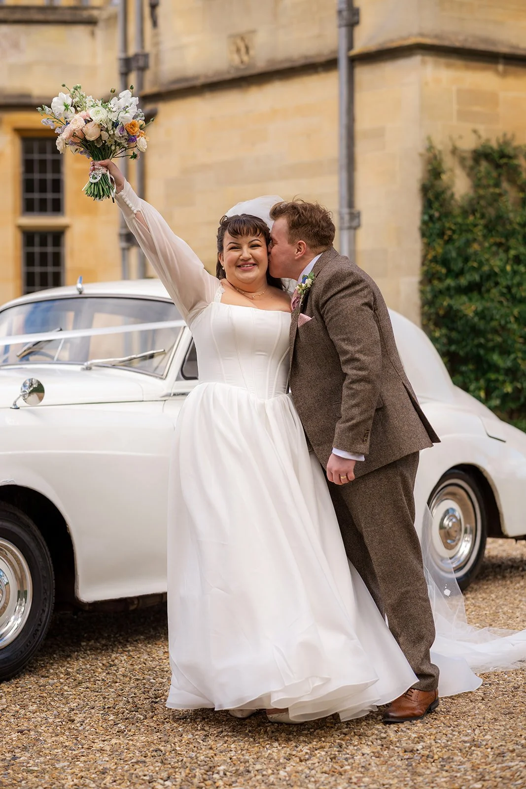 Emily & Josh stand beside a classic white wedding car at Coombe Lodge Blagdon, Somerset, UK, as Josh kisses the bride on the cheek and Emily lifts her bouquet. Wedding portrait outside the venue with soft light and elegant just-married style.