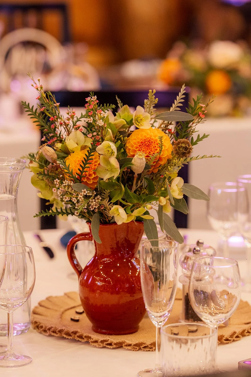 Autumn floral centrepiece at Stockbridge Farm Barn in Dorset, arranged in a rustic red jug with orange, yellow and green flowers, photographed by Just James Weddings for wedding reception detail inspiration.