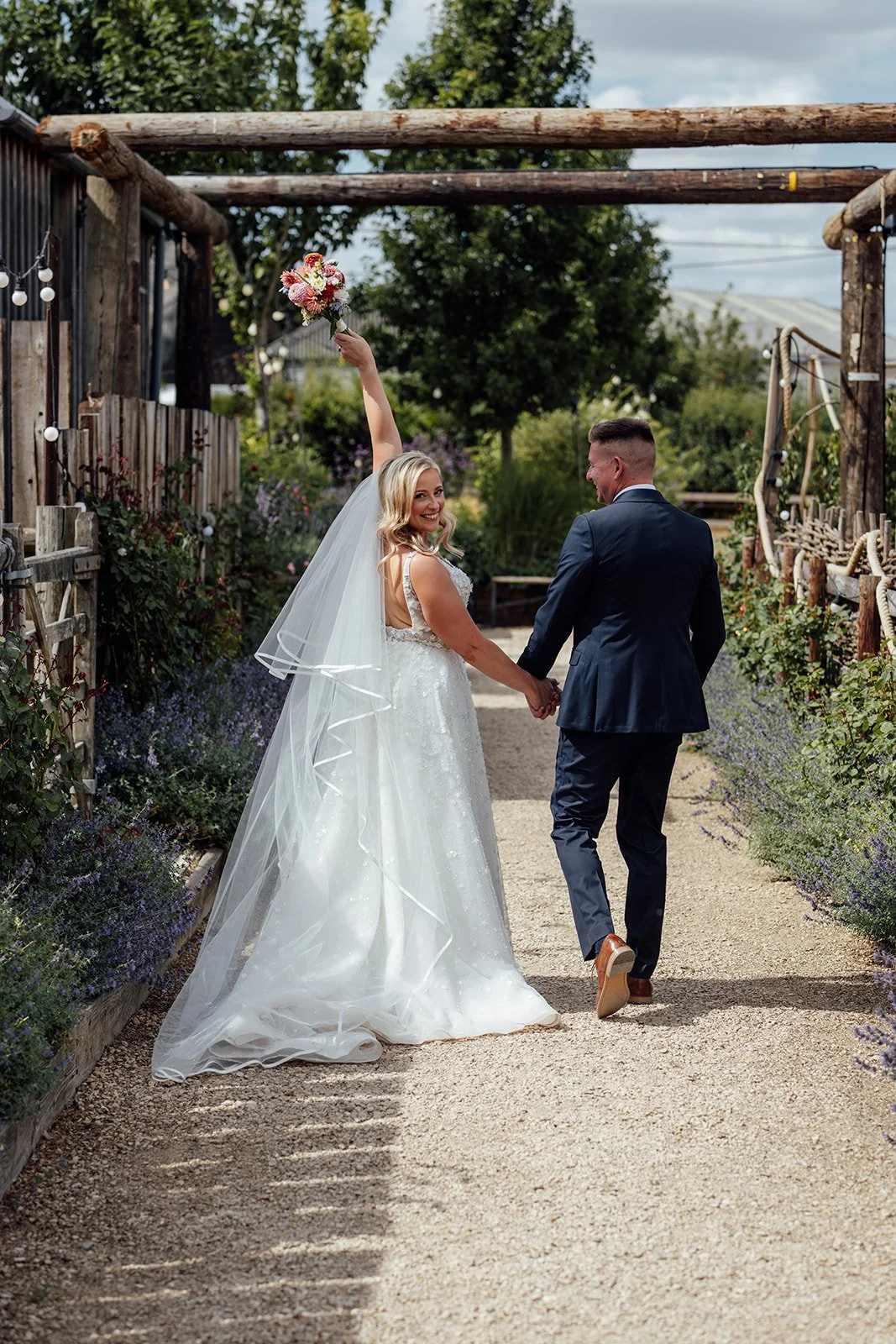 Elise looking back with her bouquet raised while walking hand in hand with Tom through the gardens at Bowerchalke Barn in Wiltshire, photographed by Just James Weddings.