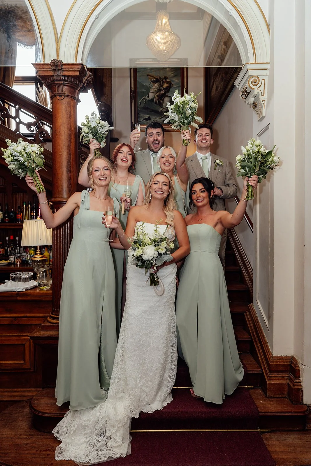 Joyful fun wedding party portrait of Amy with bridesmaids and groomsmen on the staircase inside Orchardleigh Estate in Somerset, captured by Just James Weddings with sage dresses, white bouquets, raised glasses and classic manor house.