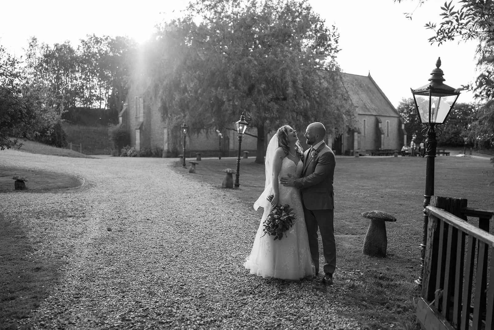 Steph &amp; Paul at Haselbury Mill sharing a quiet moment on a gravel path as evening light streams behind them. The black-and-white scene shows the couple smiling together near vintage lamps, gardens and rustic buildings.