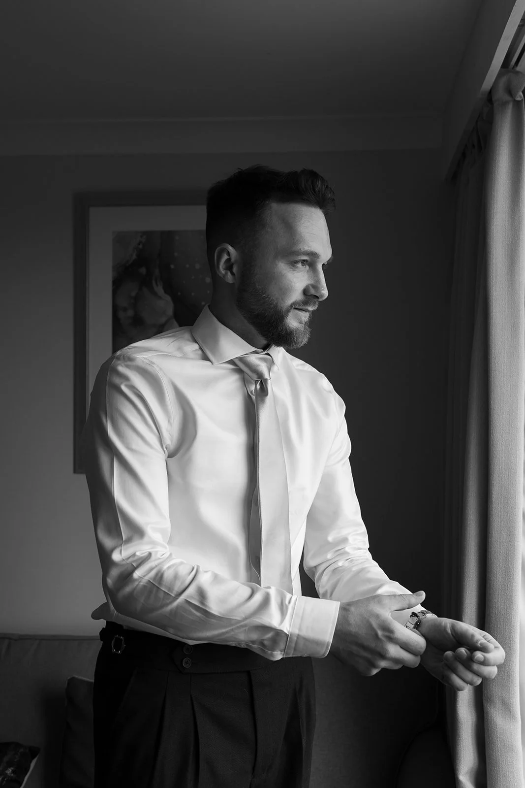 Black-and-white portrait of Connor adjusting his cufflinks beside a window while getting ready for his wedding to Amy at Sandy Cove Hotel in North Devon, UK.