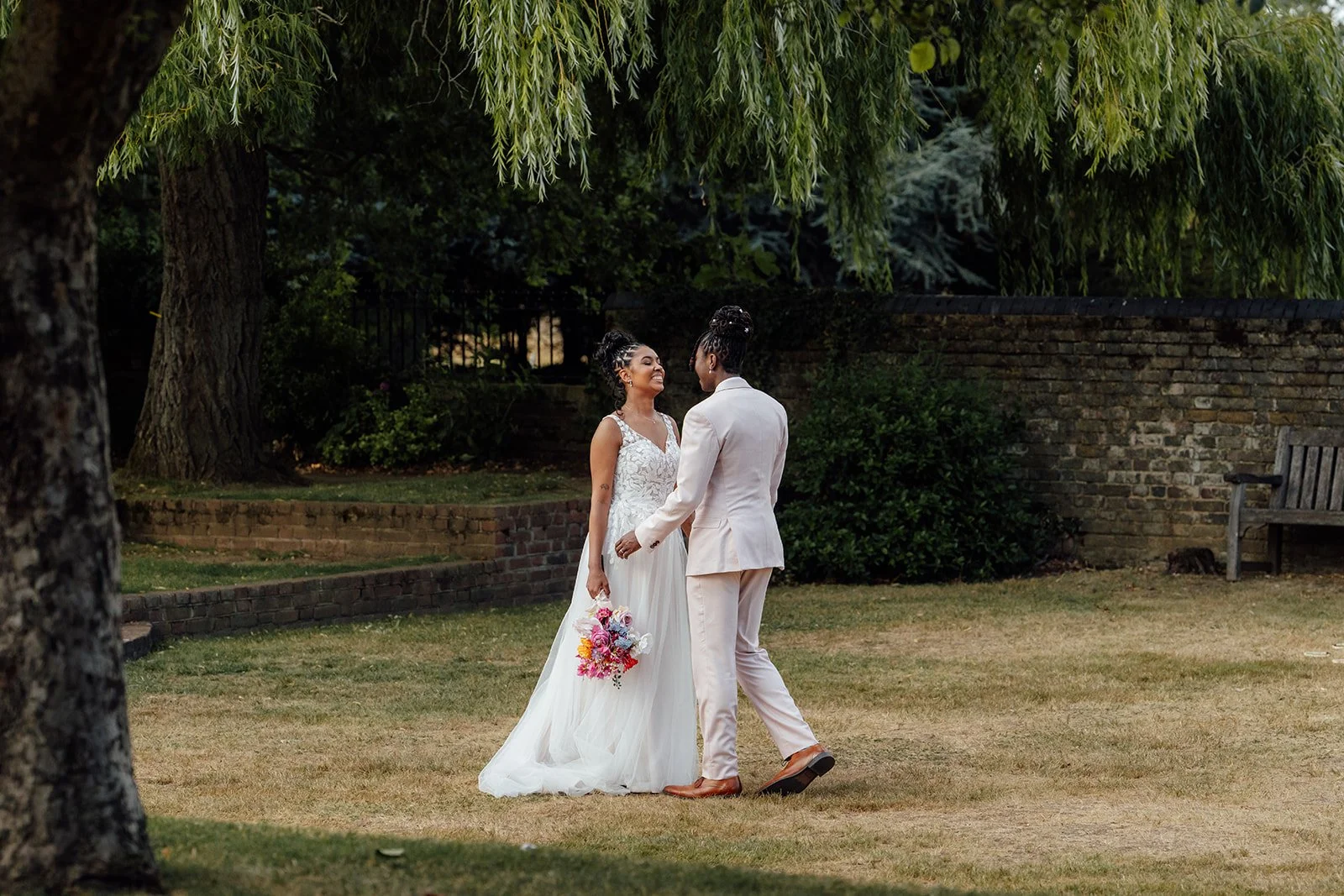Kasey & Larisha walk hand in hand beneath a large willow tree at Oaks Farm Weddings in Surrey, photographed by Just James Weddings.