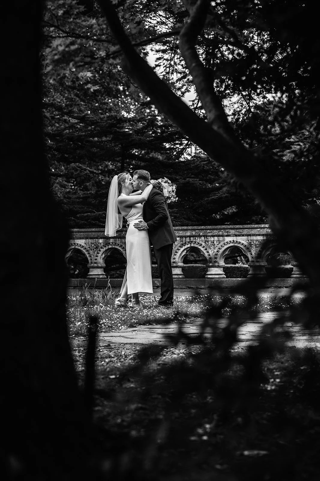 Black and white portrait of Bryony & Max kissing in the gardens at Insole Court in Cardiff, framed through dark tree branches with the decorative stone wall, soft lawn and surrounding trees behind them for a romantic outdoor wedding moment.