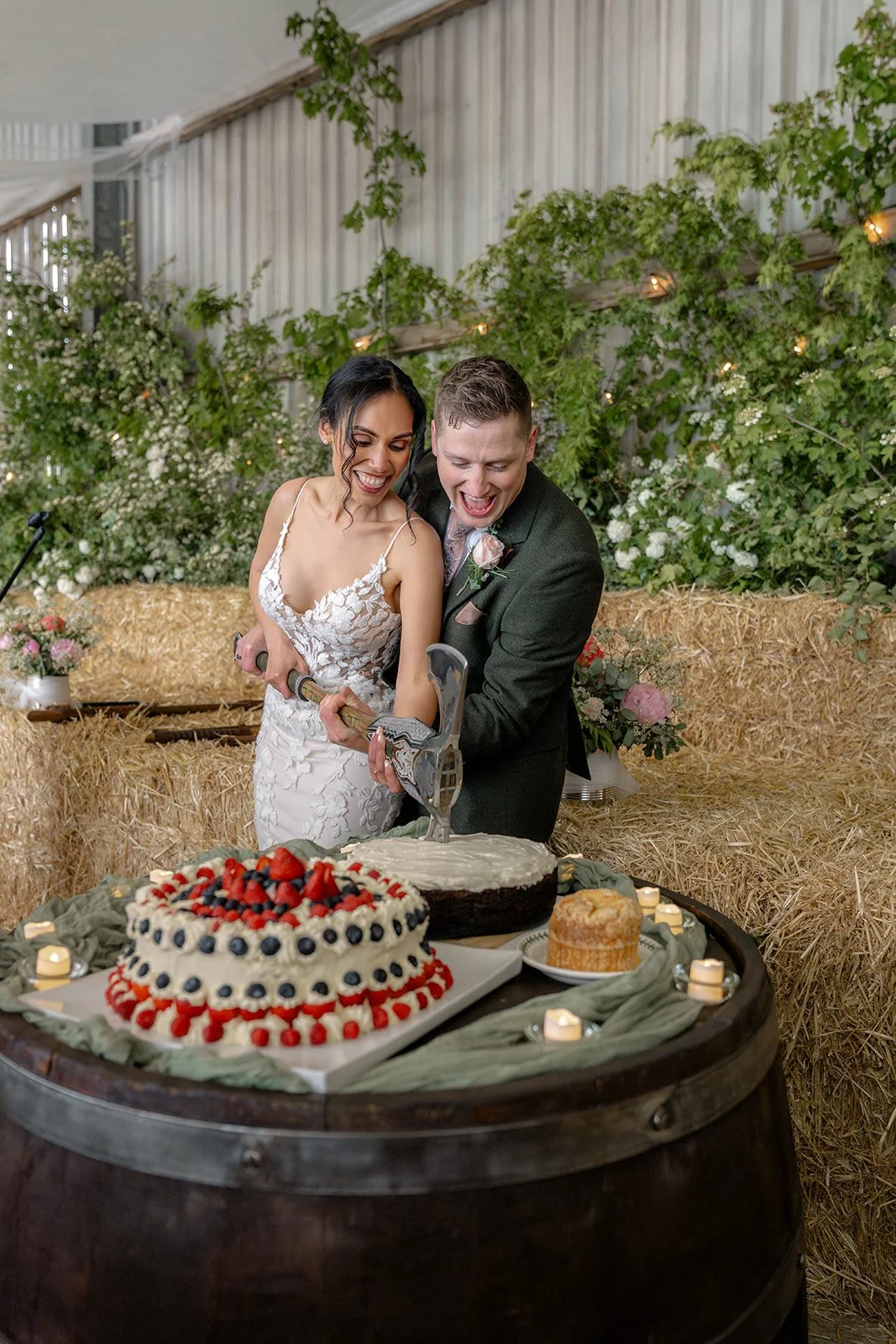 Jaye & Josh cut their wedding cake with a decorated axe at Farrington's Farm in Bristol, laughing together beside berry-topped cakes, hay bales, candlelight, greenery and relaxed rustic reception details in the barn celebration full of fun.