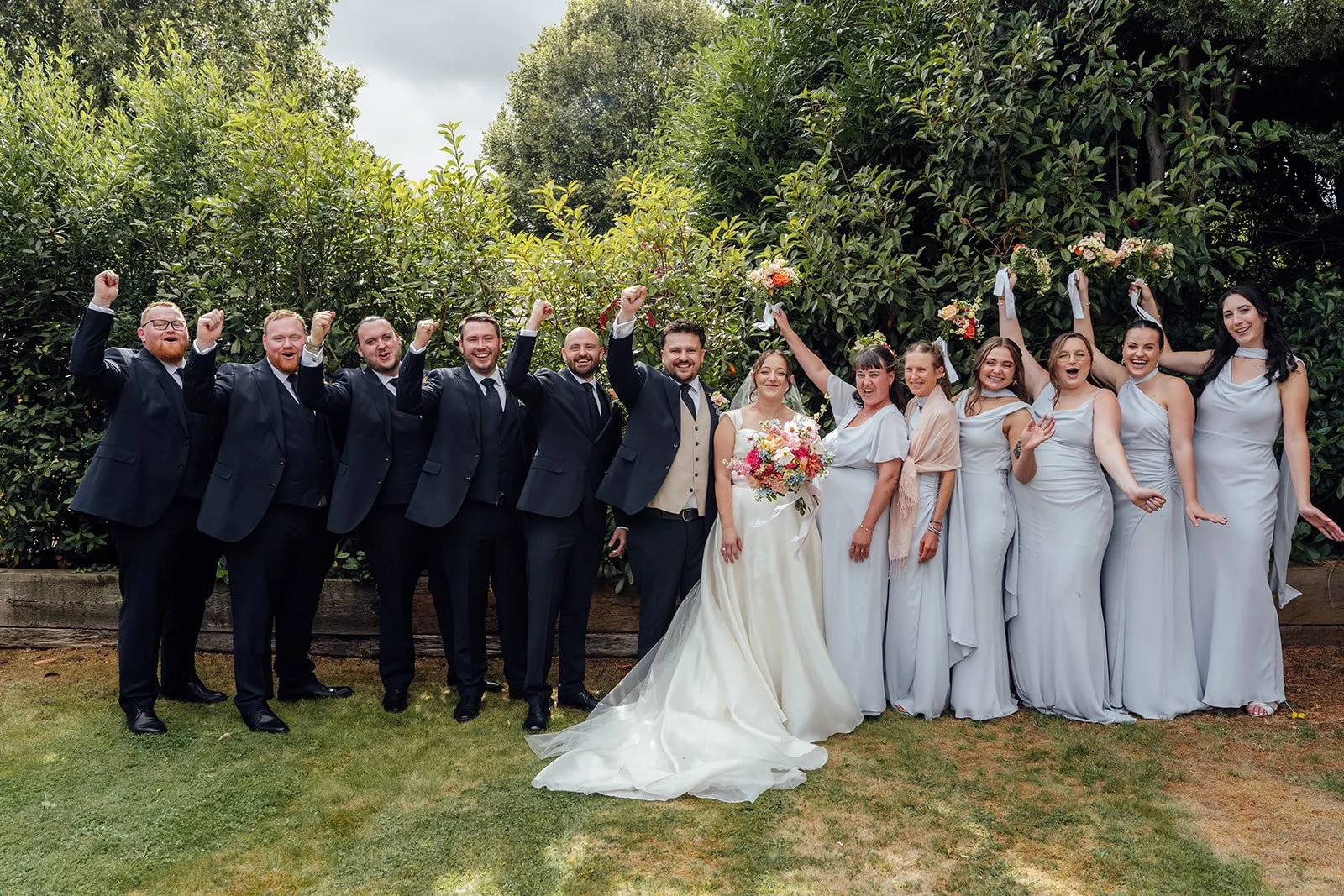Harriet & Simon pose with their full wedding party as bridesmaids raise bouquets and groomsmen cheer in the gardens at Dovecote Events in Oxfordshire, photographed by Just James Weddings.