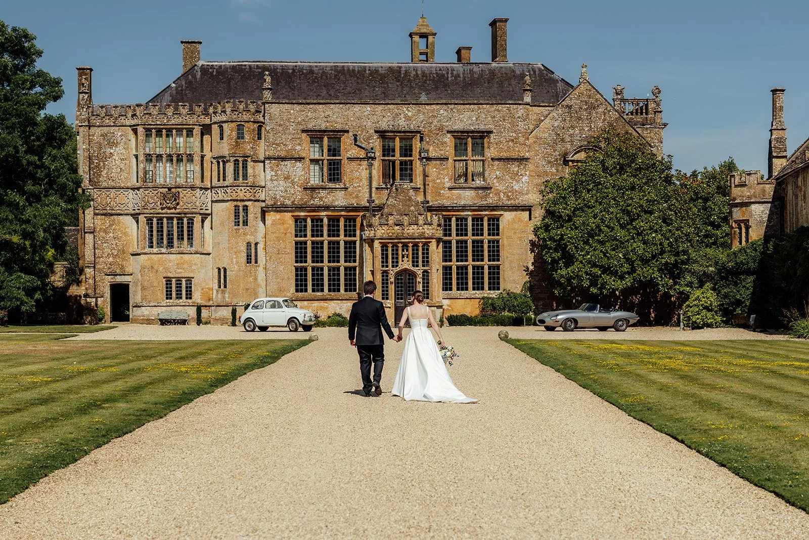 Wedding couple walking hand in hand along the gravel driveway towards Brympton House in Somerset, with the historic manor and classic cars behind them, photographed by Just James Weddings.
