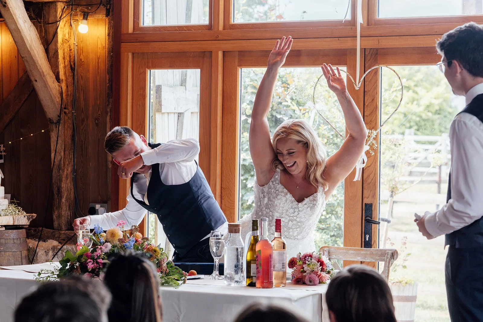 Elise celebrating with her arms raised beside Tom during the wedding speeches inside Bowerchalke Barn in Wiltshire, photographed by Just James Weddings.