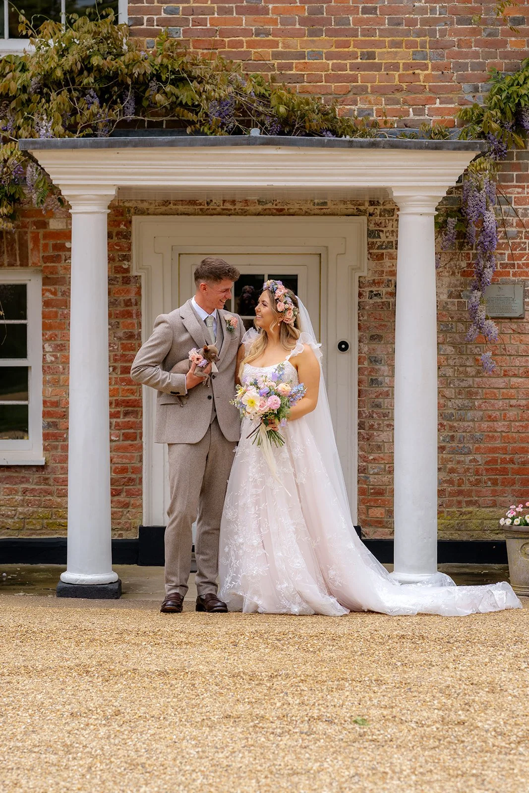 Jaye & Josh standing together outside Syrencot in Wiltshire, framed by white columns and brick walls, photographed by Just James Weddings for relaxed wedding portraits.
