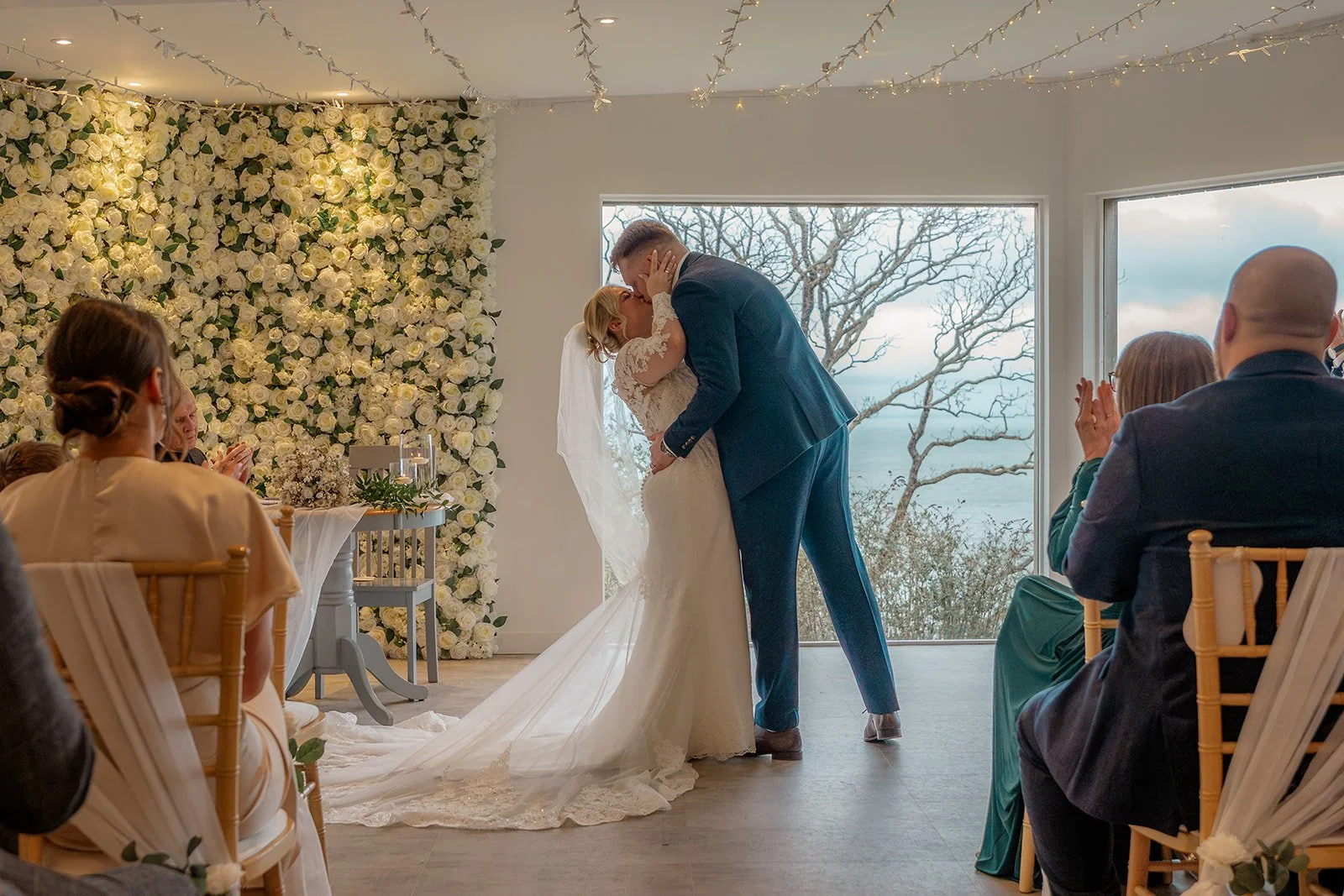 Daisy and Andrew share their first kiss as their guests rise and applaud during their ceremony at Sandy Cove Hotel in Devon, in front of a lush white rose flower wall with large coastal windows framing the sea and bare winter trees behind them.