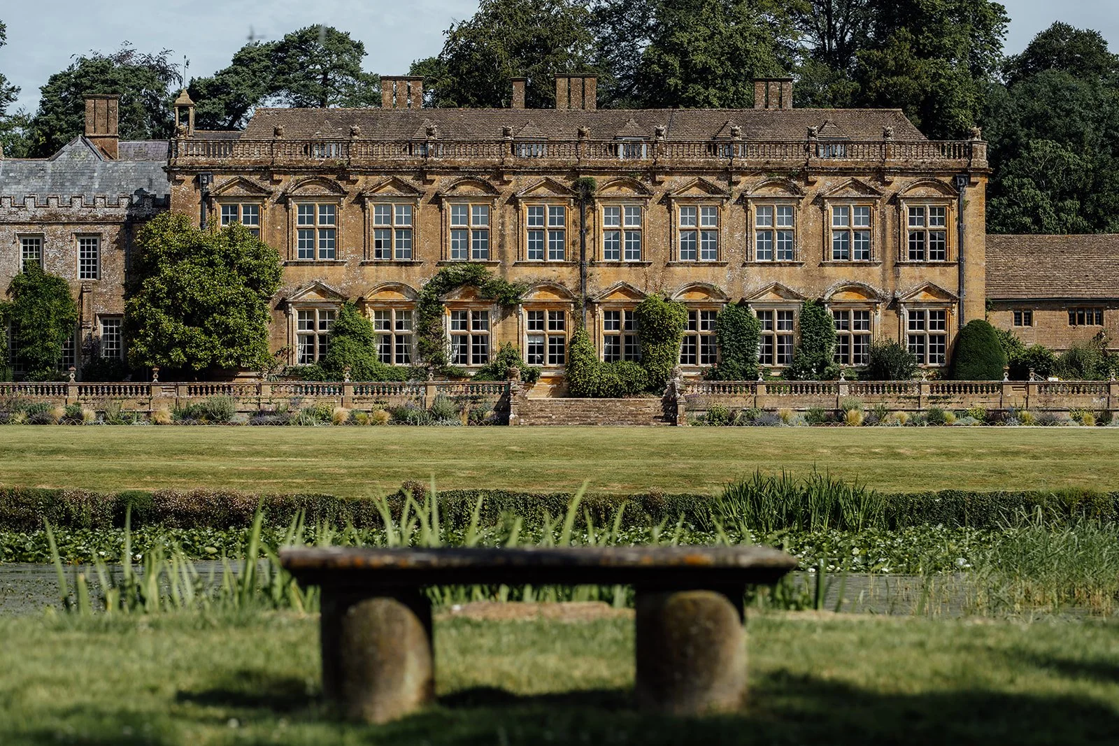 View of Brympton House in Somerset from the gardens, with the historic manor, formal lawn and stone bench in the foreground, photographed by Just James Weddings.