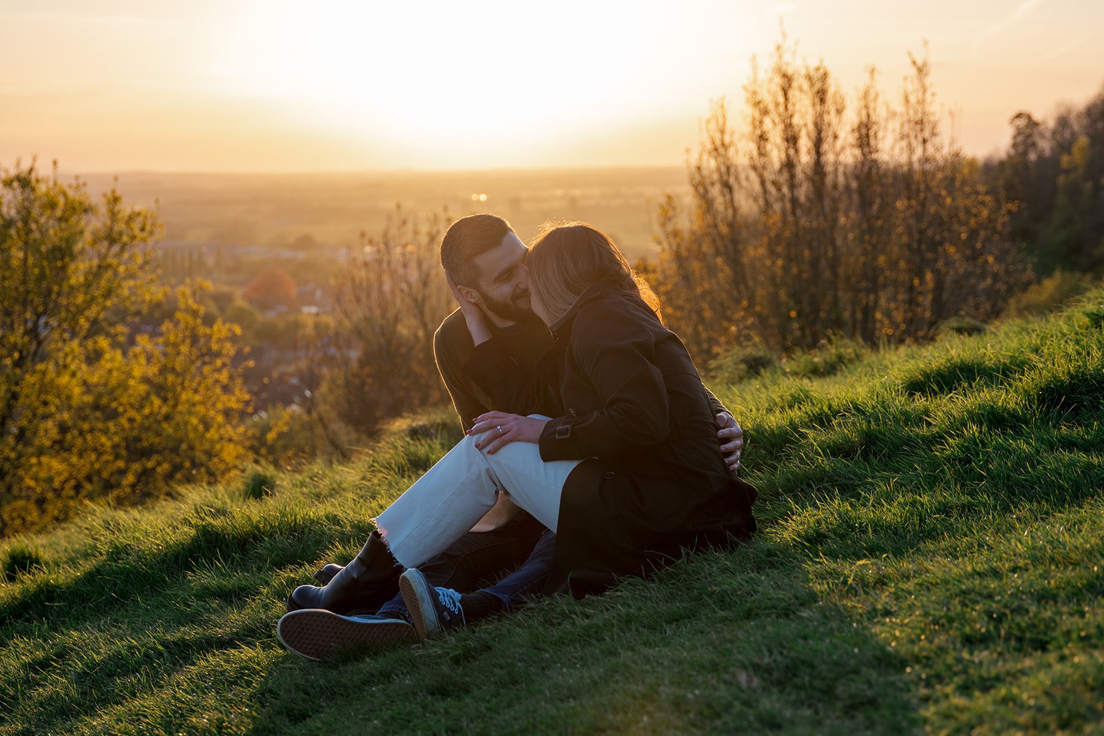 Roy and Julia sitting close together on a grassy Somerset hillside at sunset, sharing a quiet kiss with glowing countryside views and soft golden evening light behind them during a relaxed engagement shoot photographed by Just James Weddings.