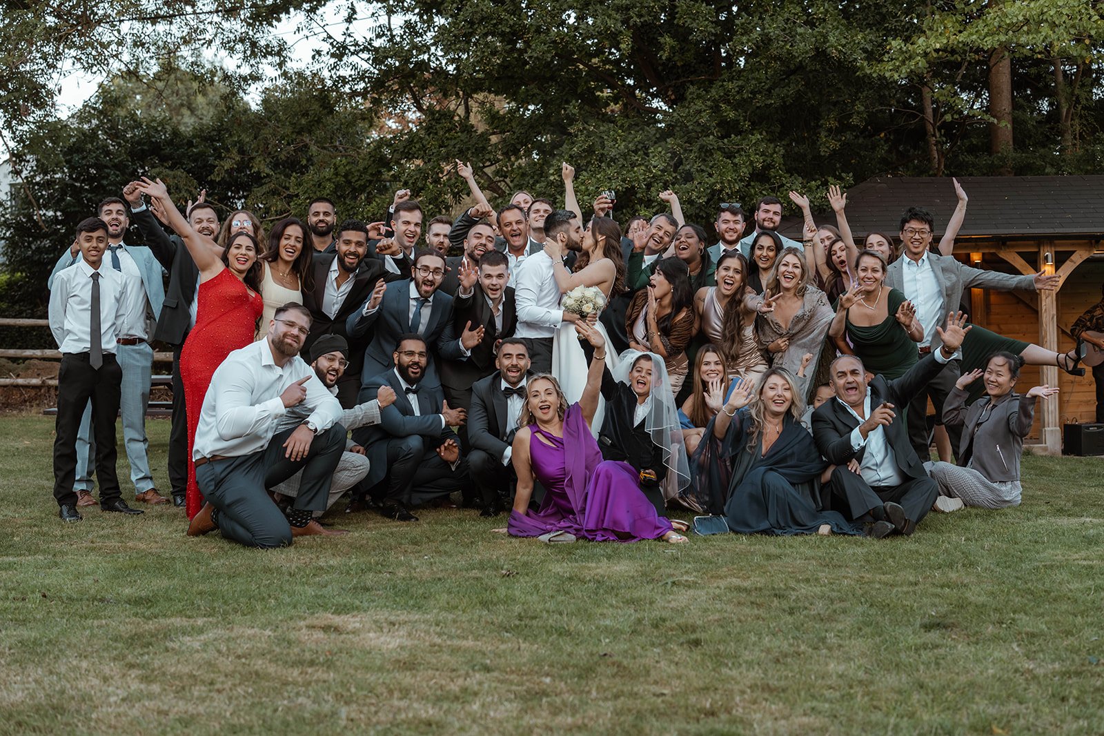 Roy &amp; Julia at The Crown Inn celebrating with a large group of guests outdoors. The couple kiss at the centre while everyone cheers, poses and raises their hands, creating a joyful, energetic group photo on the lawn with trees in the background.