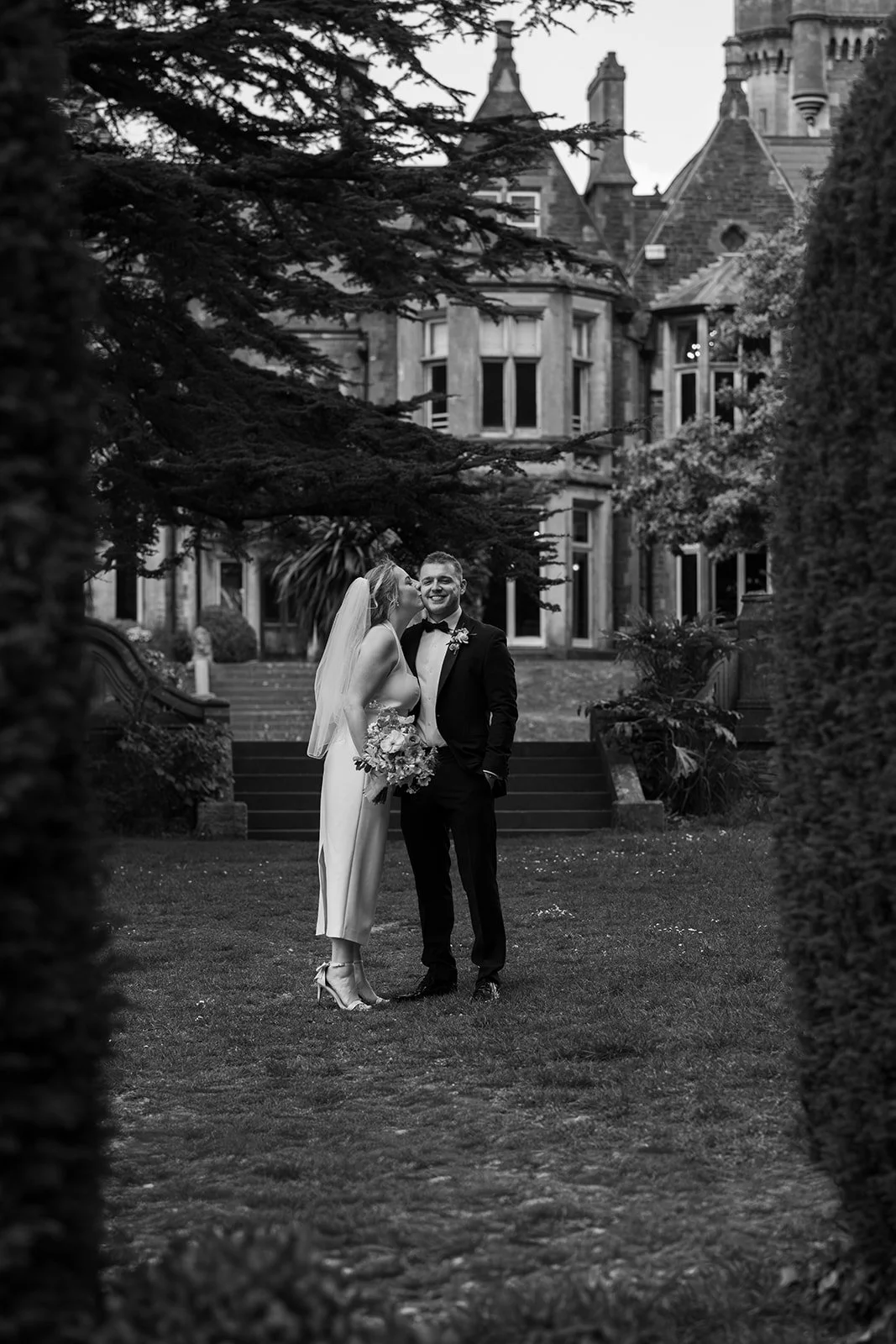 Black and white portrait of Bryony & Max in the gardens at Insole Court in Cardiff, with Bryony kissing Max’s cheek as he smiles, framed by trees, lawn, hedges and the historic house behind them for a relaxed outdoor wedding portrait together.