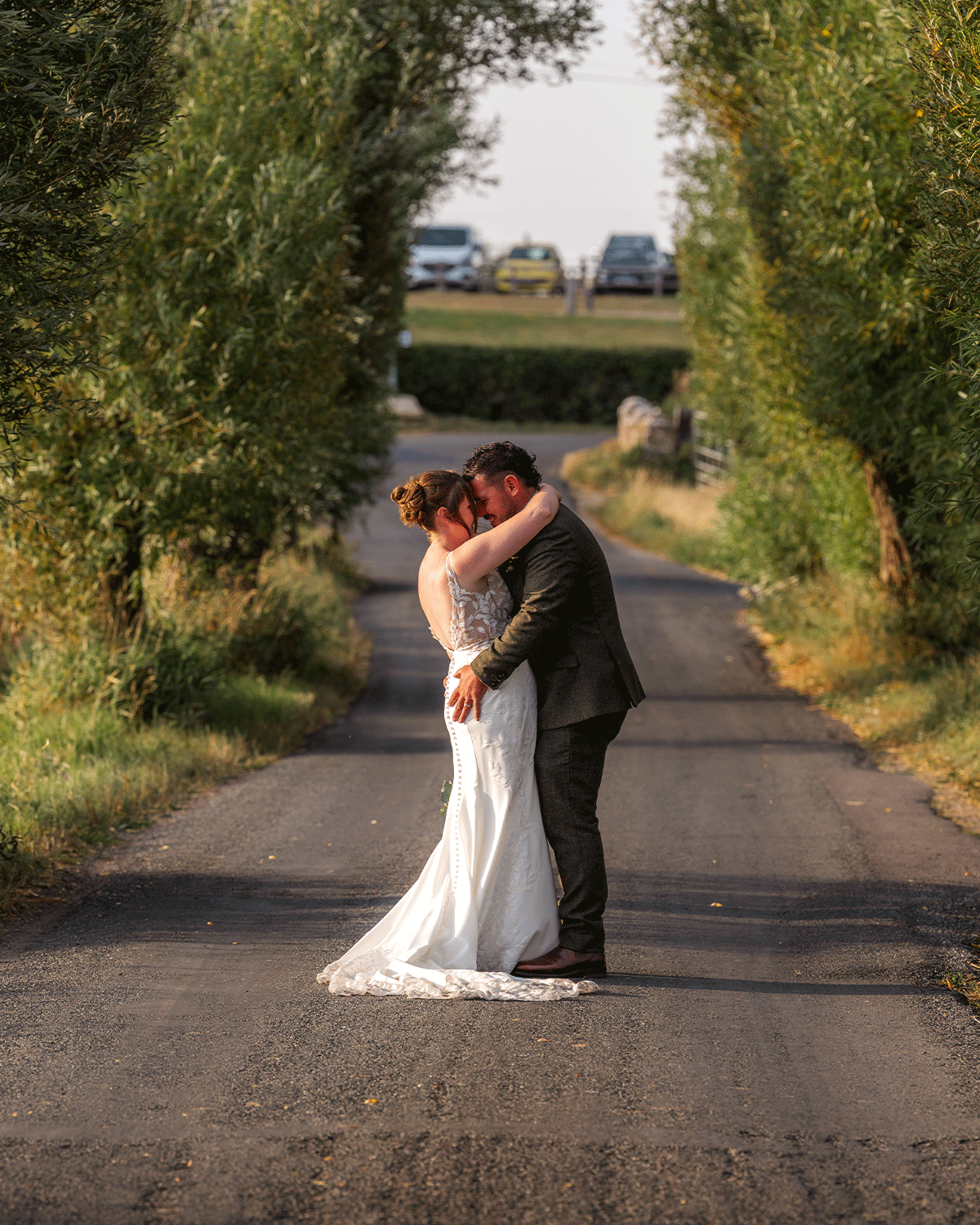 Shannon & Lewis kissing in the middle of a quiet country road in Glastonbury, somerset, on their wedding day. The bride’s lace gown trails behind her as the groom holds her close, with tall hedges, parked cars, and warm evening light.
