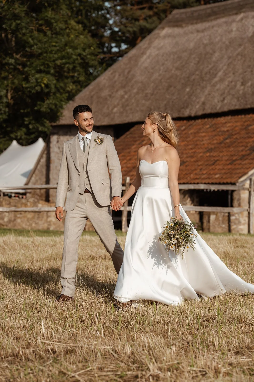 Louise & Christian walking through the field at The Cider Barn in Somerset, with rustic barn buildings behind them, photographed by Just James Weddings for relaxed countryside wedding portraits.
