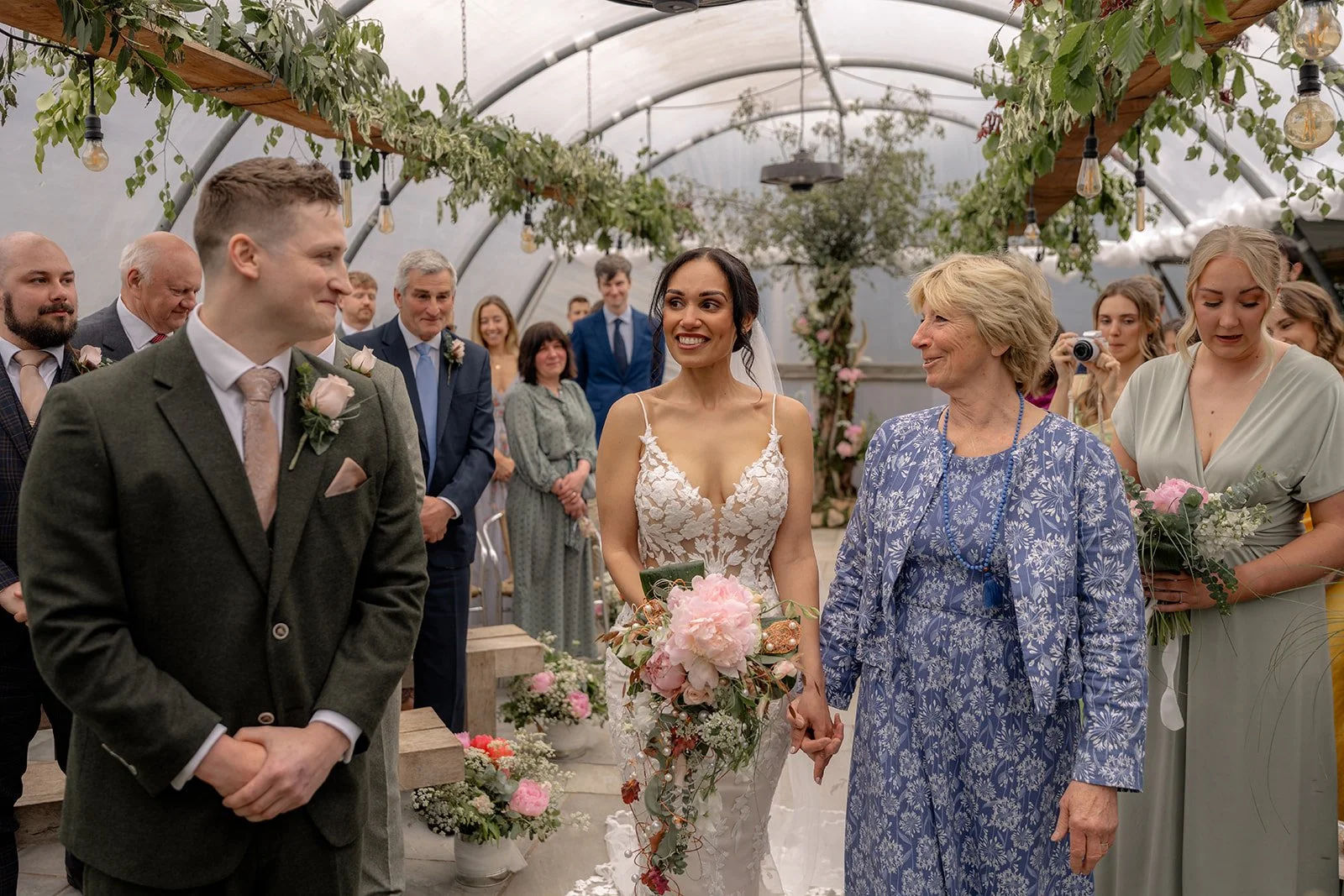 Jaye & Josh at Farrington's Farm in Bristol, with the bride walking down the aisle beside her mum as the groom smiles ahead, surrounded by guests, bridesmaids, hanging greenery, soft pink wedding flowers and a relaxed poly tunnel ceremony.