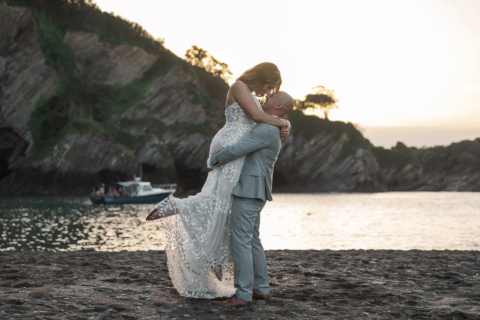 Toya & Simon sharing a sunset kiss on the beach at Sandy Cove Hotel in North Devon, with the groom lifting the bride beside the sea and cliffs, photographed by Just James Weddings for romantic coastal wedding portraits in golden evening light.