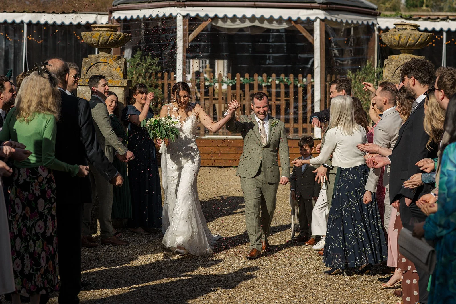 Anna & Chris walk through confetti outside Elmhay Park in Somerset, UK, hand in hand as guests cheer around them in the afternoon sunshine. A lively, joyful wedding moment from Anna & Chris’s day, filled with celebration, movement, and love.