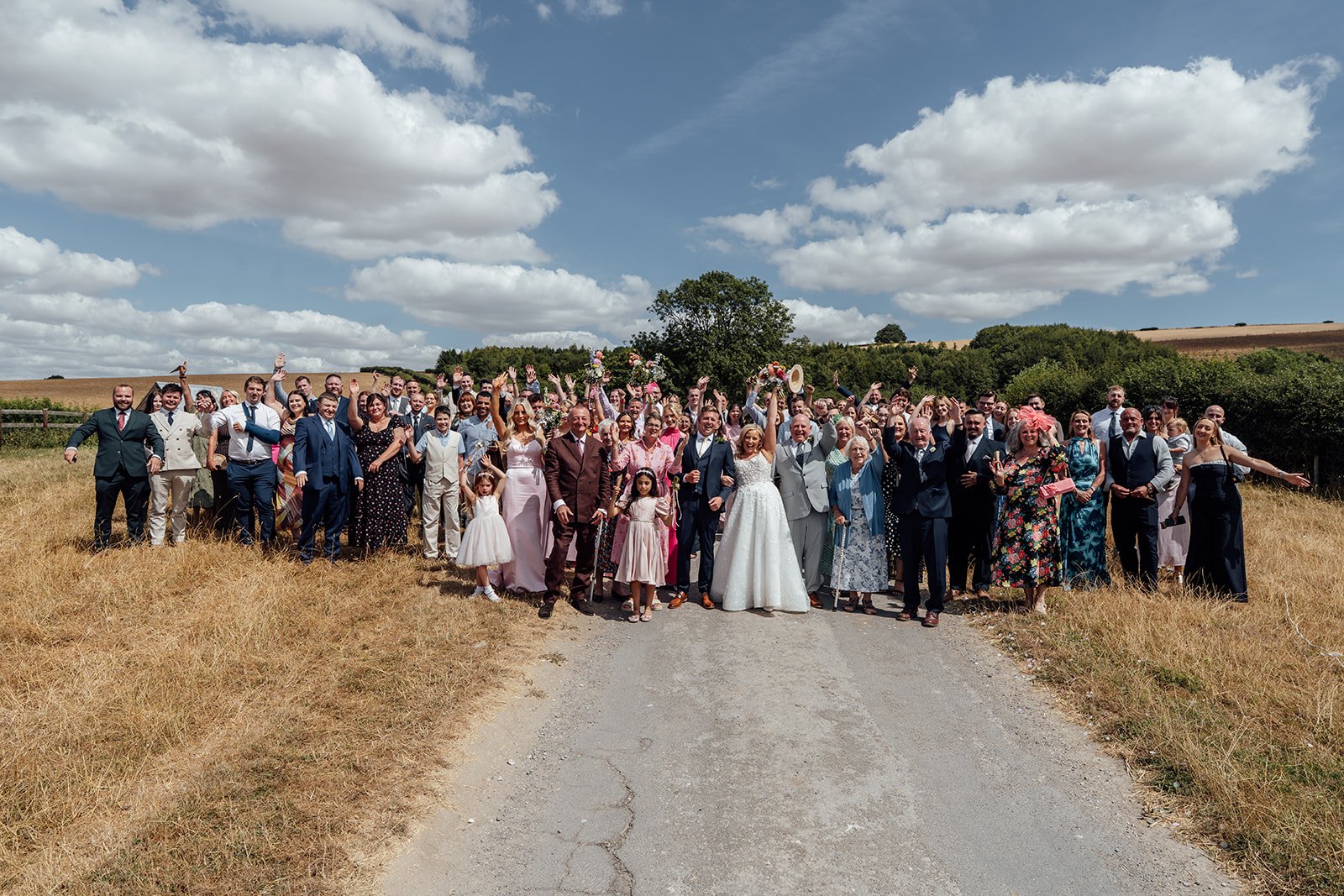 Elise and Tom standing with their wedding guests for a large countryside group photo at Bowerchalke Barn in Wiltshire, photographed by Just James Weddings.