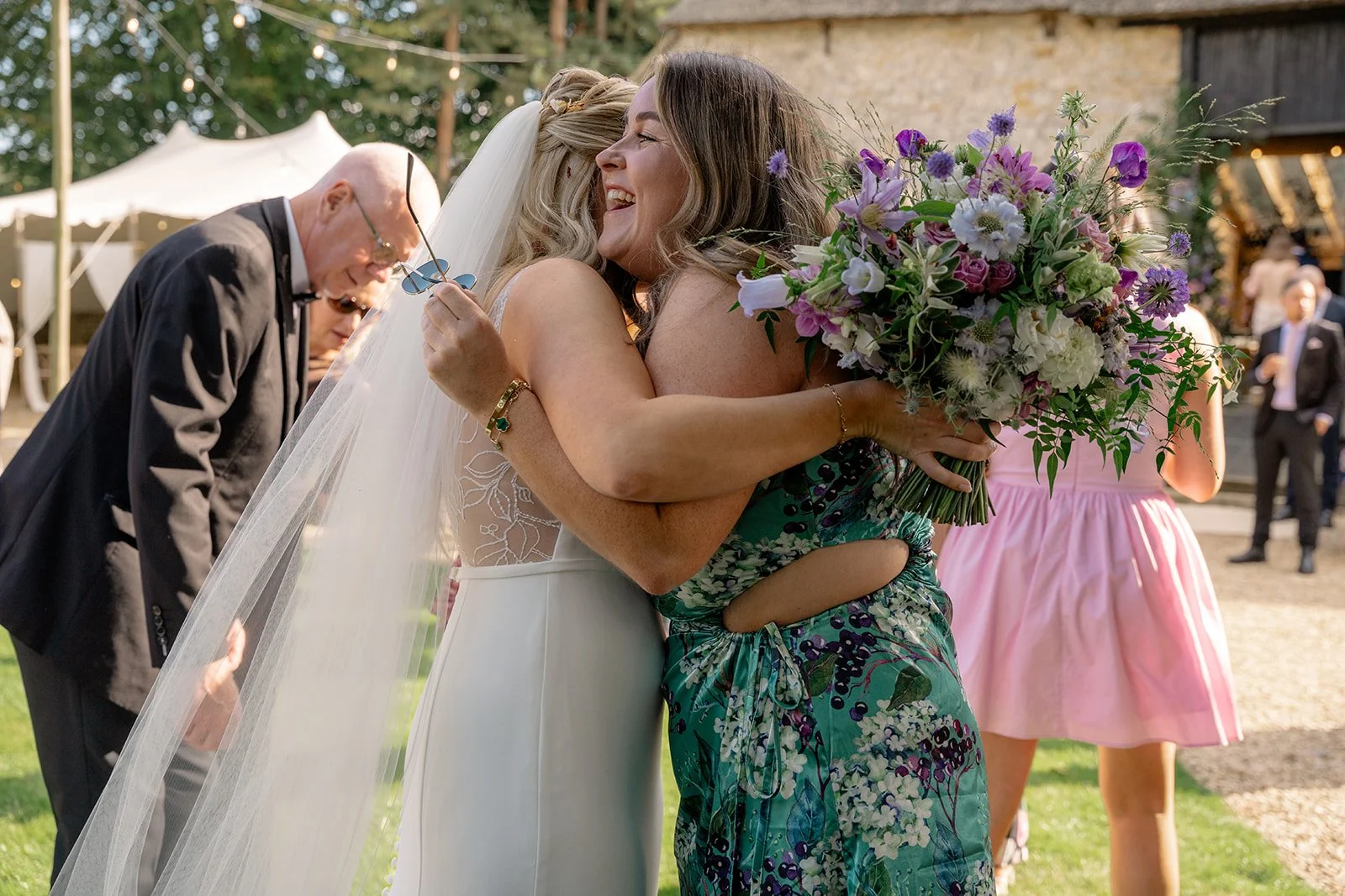 Abi hugging a guest after the ceremony at The Cider Barn in Somerset, holding a colourful bouquet and laughing outdoors, photographed by Just James Weddings for joyful documentary wedding moments.