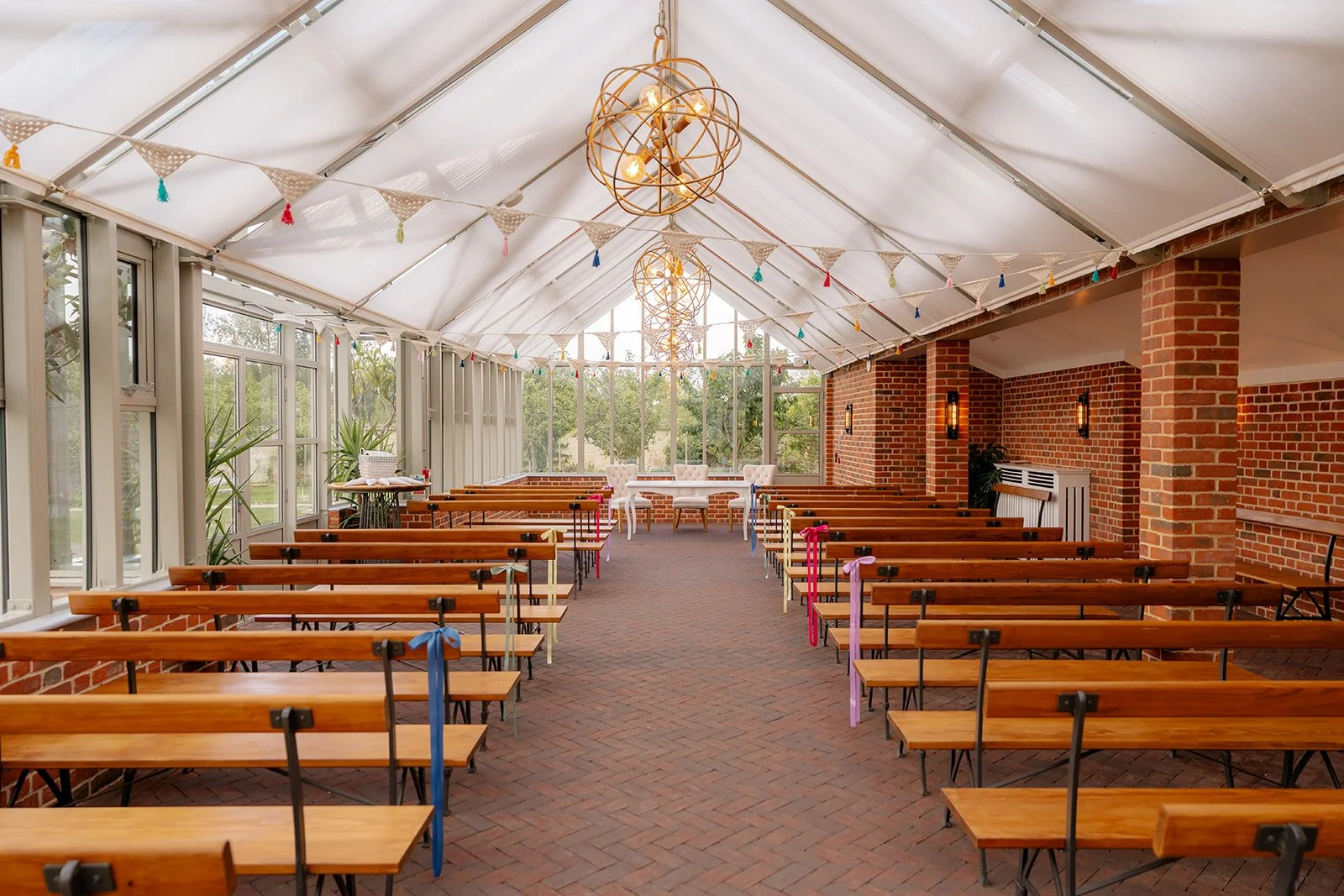 Indoor ceremony space at Syrencot in Wiltshire, with wooden benches, bunting and pendant lights beneath a bright glass roof, photographed by Just James Weddings for wedding venue inspiration.