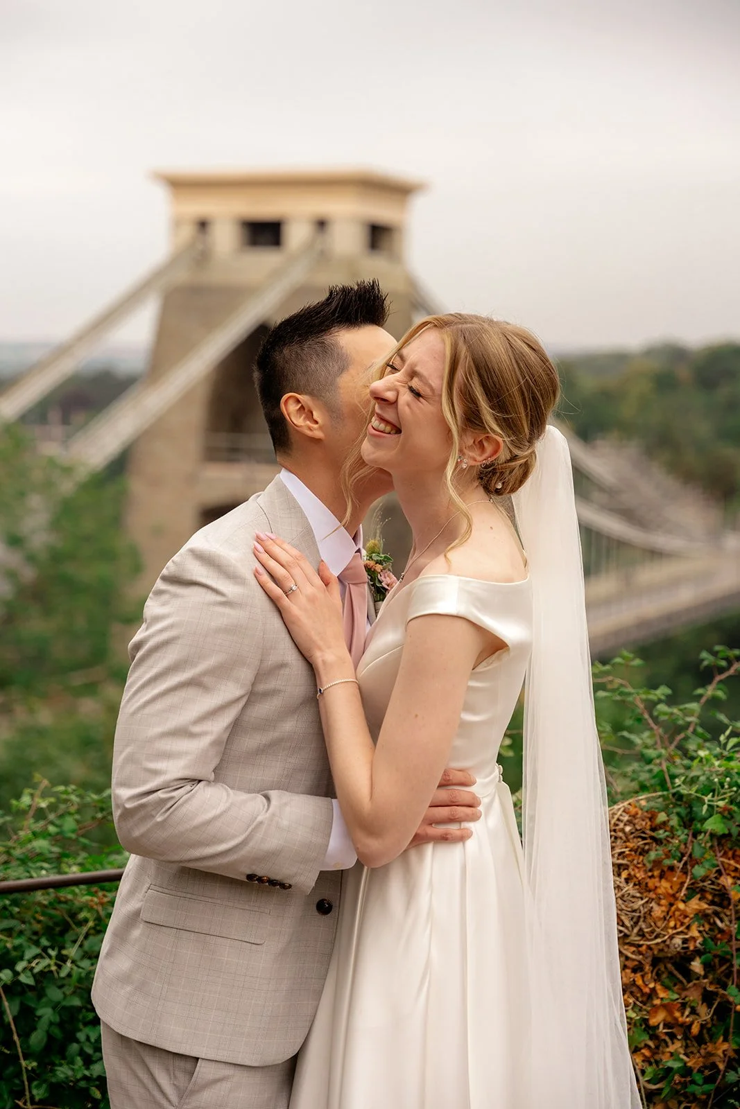 Brian & Josie laughing together at Clifton Observatory in Somerset, with the Clifton Suspension Bridge behind them, photographed by Just James Weddings for relaxed wedding portraits.