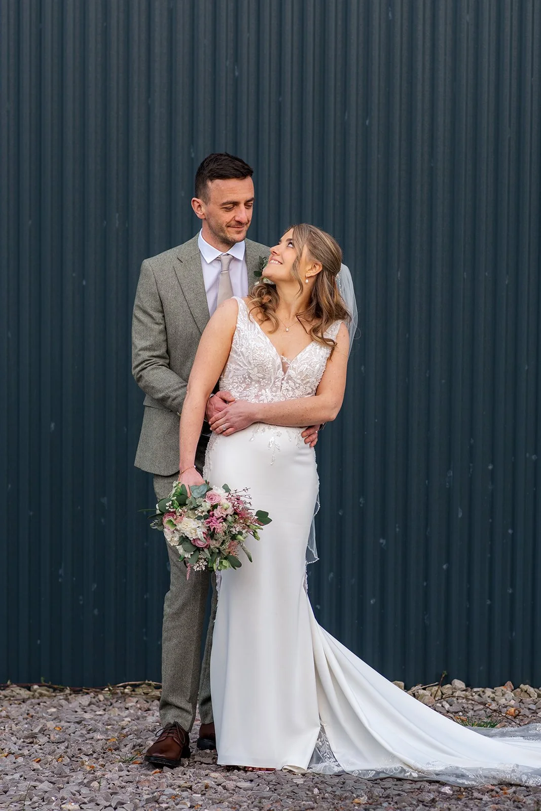 Anna and Tom pose for portraits at Winkworth Farm in the Cotswolds, UK, with Tom standing behind Anna as she holds her bouquet. This elegant couple portrait features Anna’s lace gown and a dark barn backdrop in Cotswolds wedding photography.