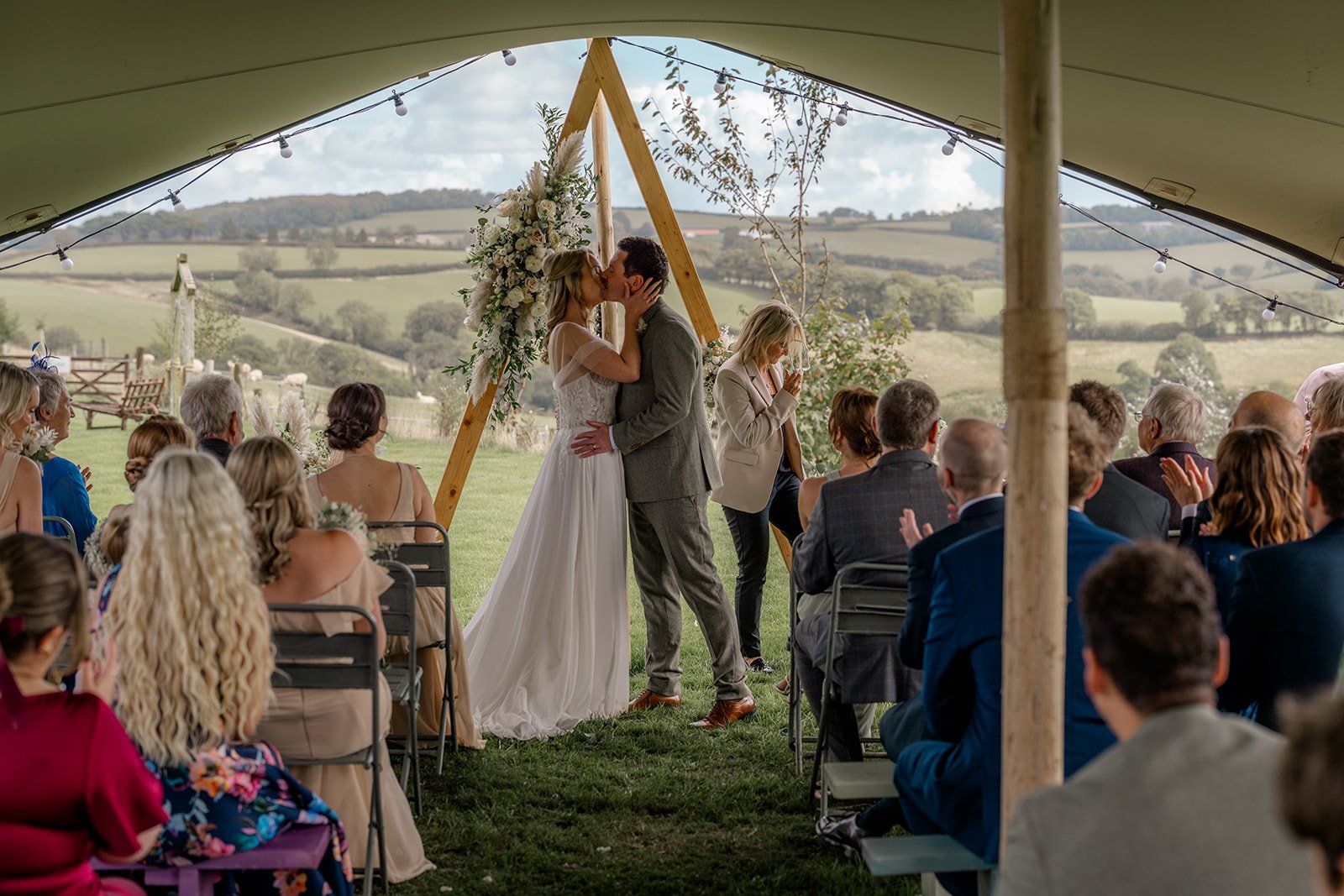 Samantha & Mike sharing their first kiss during their outdoor wedding ceremony at The Hawke Barn, devon. Guests sit beneath a canvas canopy as rolling countryside stretches behind them, with floral arrangements and soft light framing the moment.