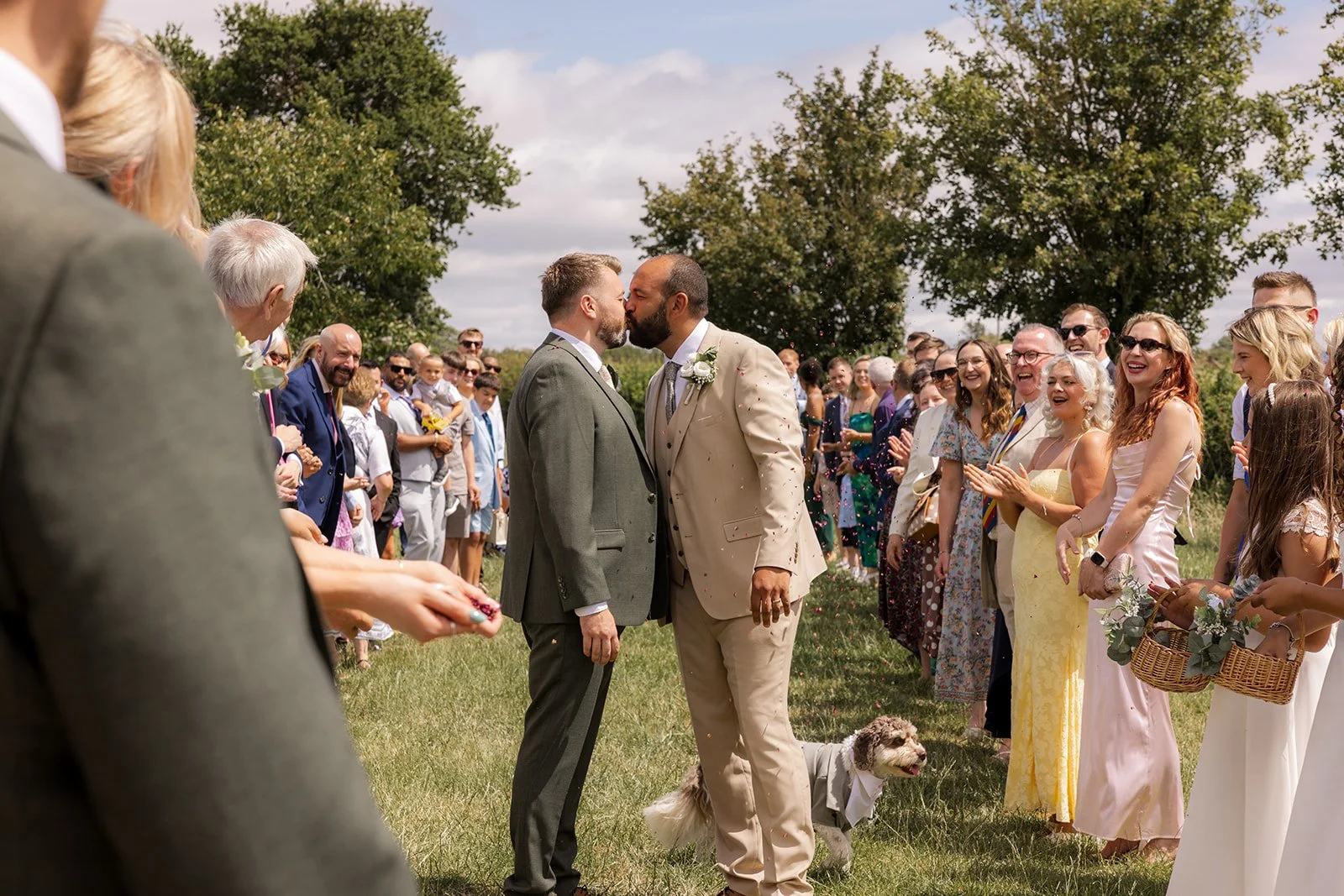 Aaron & Jack pause for a kiss with Milo beside them as smiling guests throw colourful confetti outside Rock Village Hall in Worcestershire, photographed by Just James Weddings.