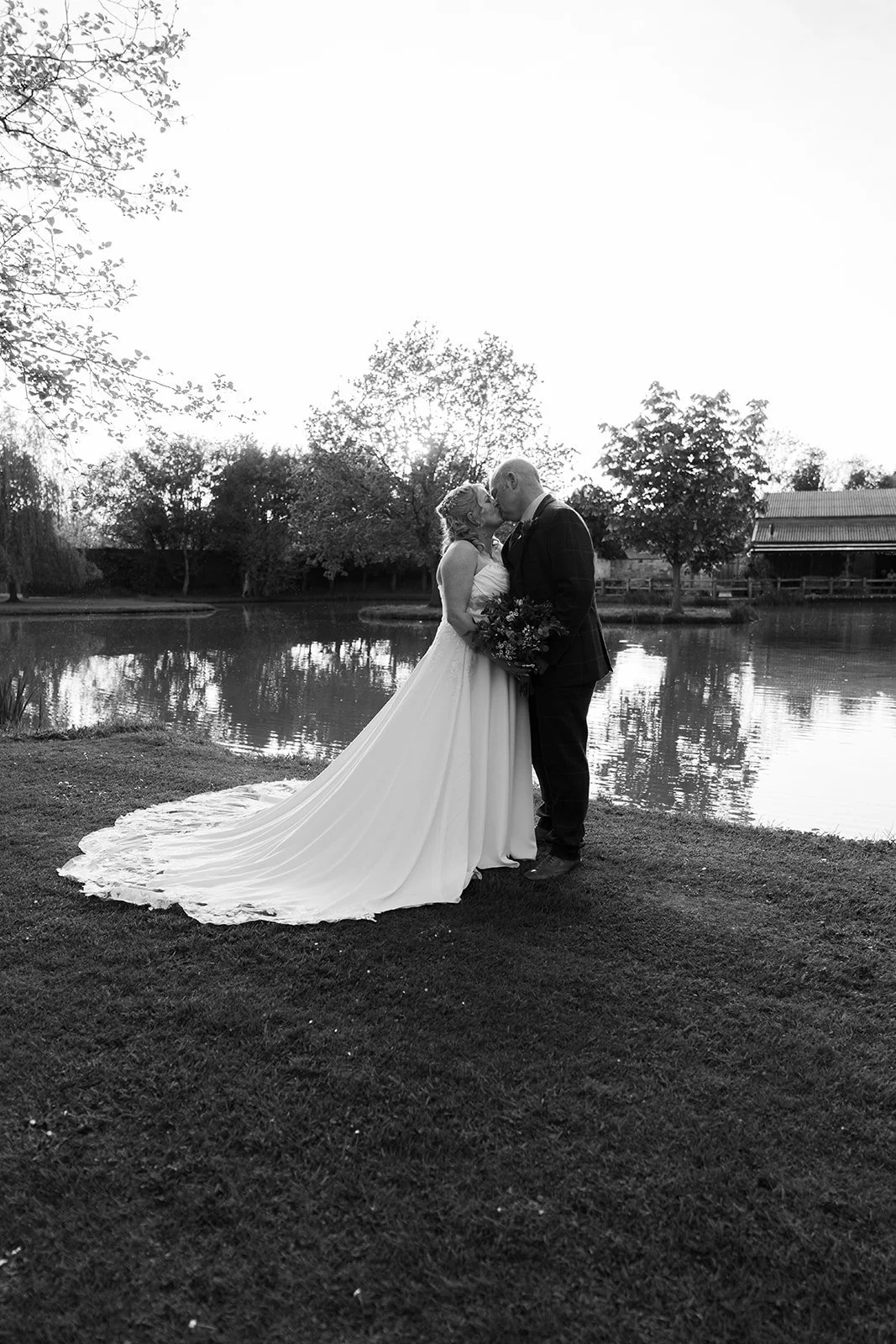 Debbie and Nigel kiss beside the lake at Yarlington Barn in Somerset in a black and white wedding portrait, with Debbie’s dress flowing across the grass and reflections on the water adding a timeless countryside feel.