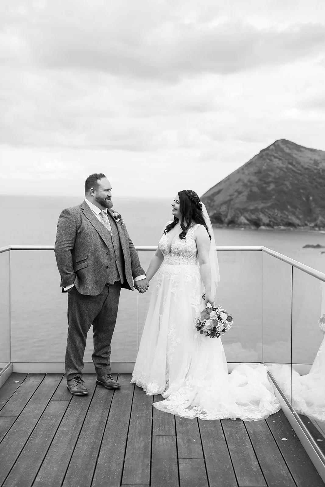 Black and white portrait of Sam & Terri holding hands on the balcony at Sandy Cove Hotel in North Devon, with sea cliffs behind them, photographed by Just James Weddings for relaxed coastal wedding portraits with natural scenery and calm views.