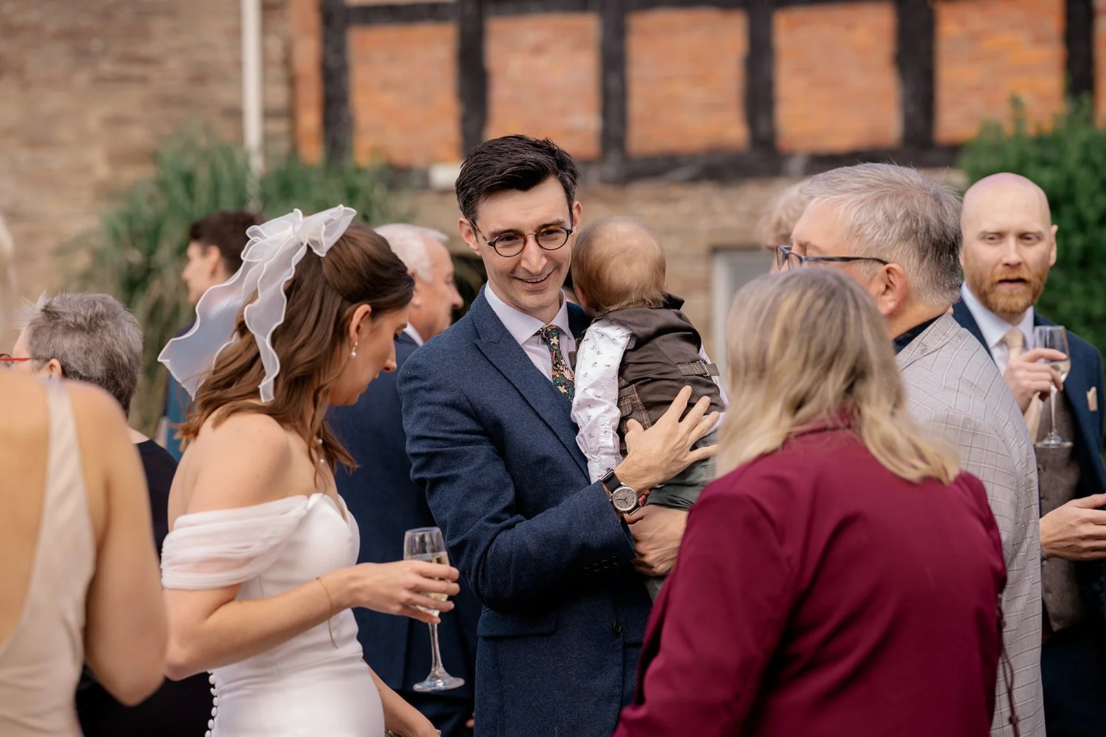 Lloyd holding a baby while chatting with guests during the drinks reception at Broadfield Court in Herefordshire, photographed by Just James Weddings for candid wedding moments.