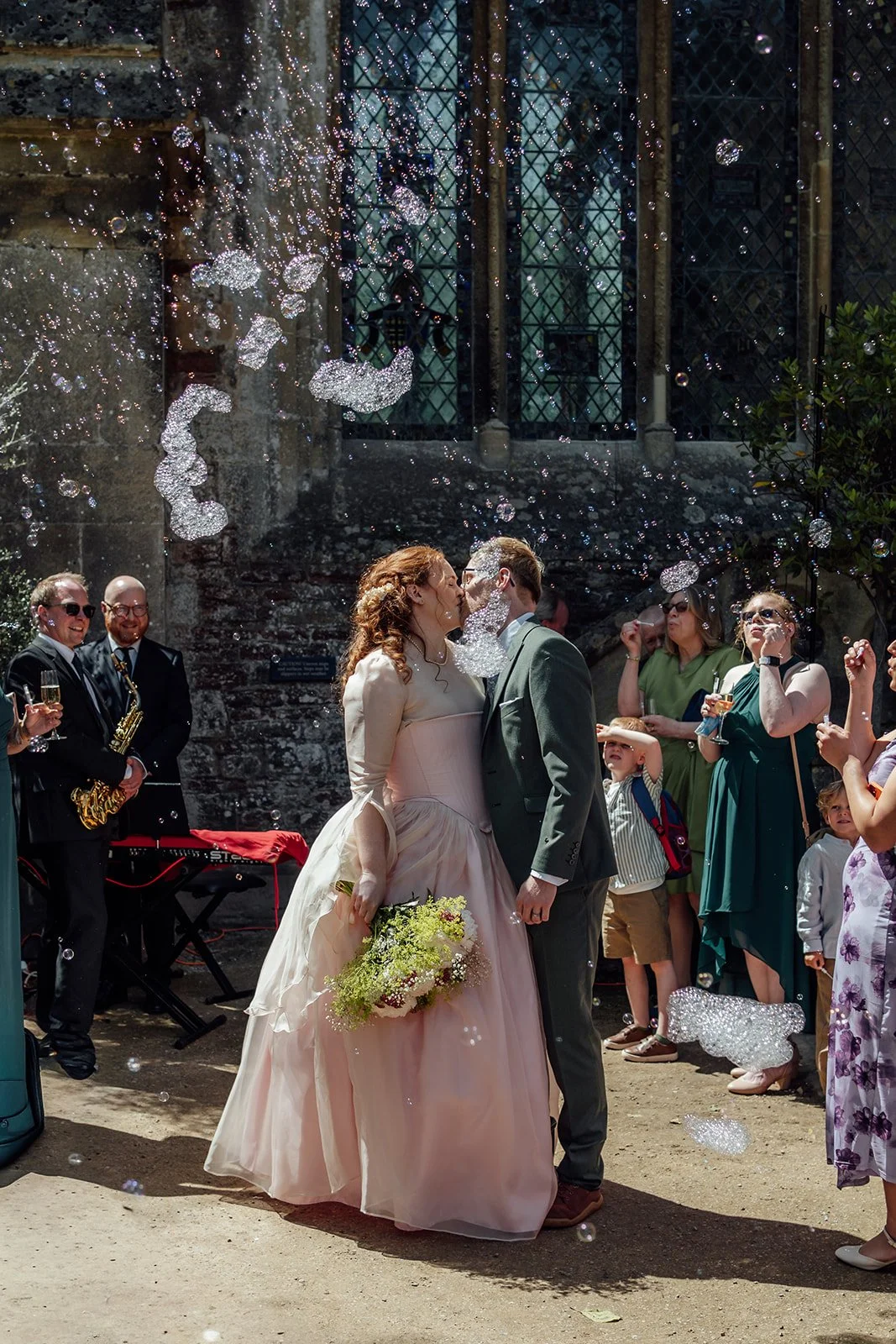 Charlotte & Jack kiss at Bishop’s Palace in Somerset, UK, surrounded by bubbles, guests and live musicians in the sunny courtyard, with the bride holding her bouquet and the historic stained glass window behind them during reception drinks.