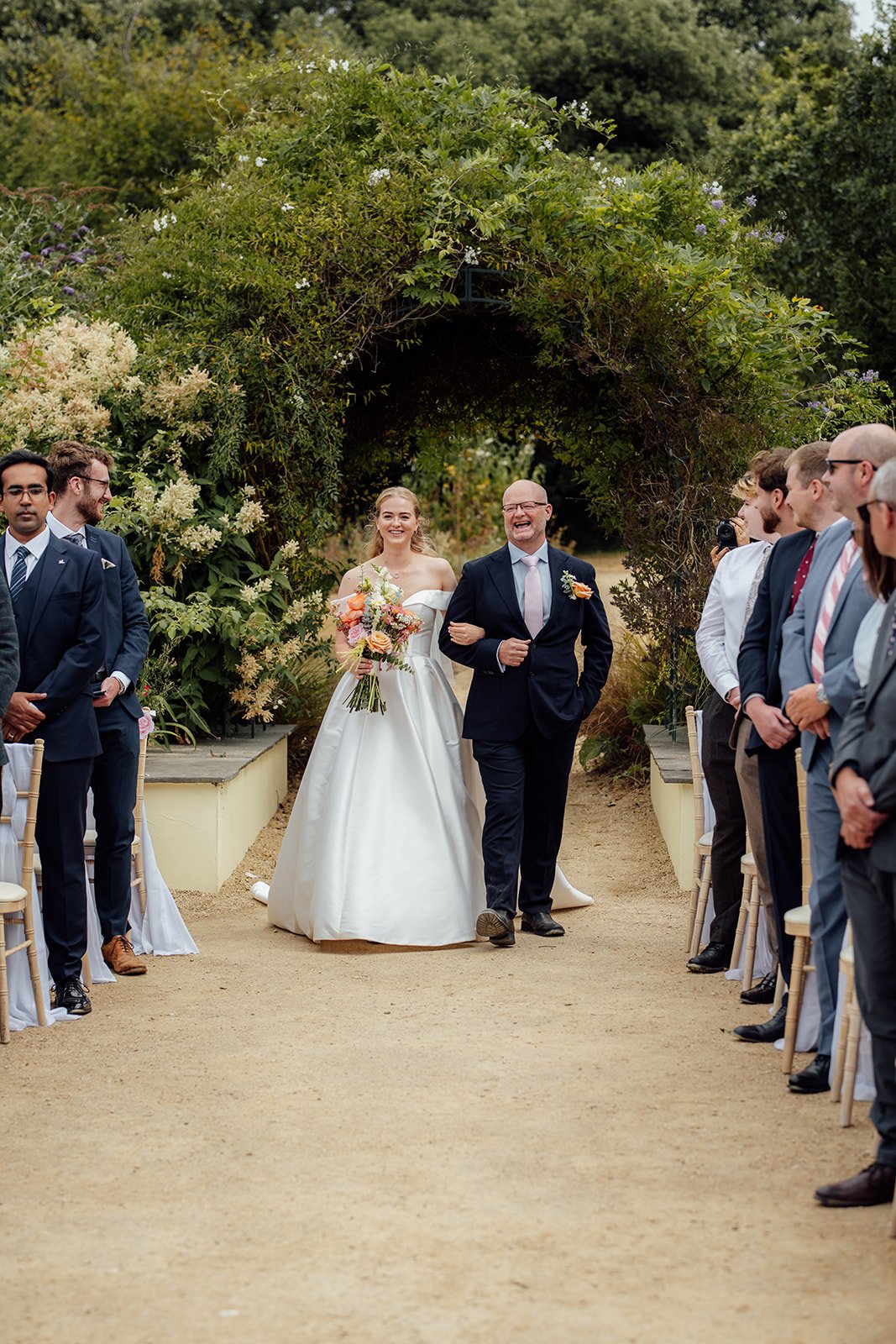 Caitlin walking down the outdoor ceremony aisle with her father at Clevedon Hall in Somerset, carrying a colourful bouquet as guests watch beneath the garden archway.