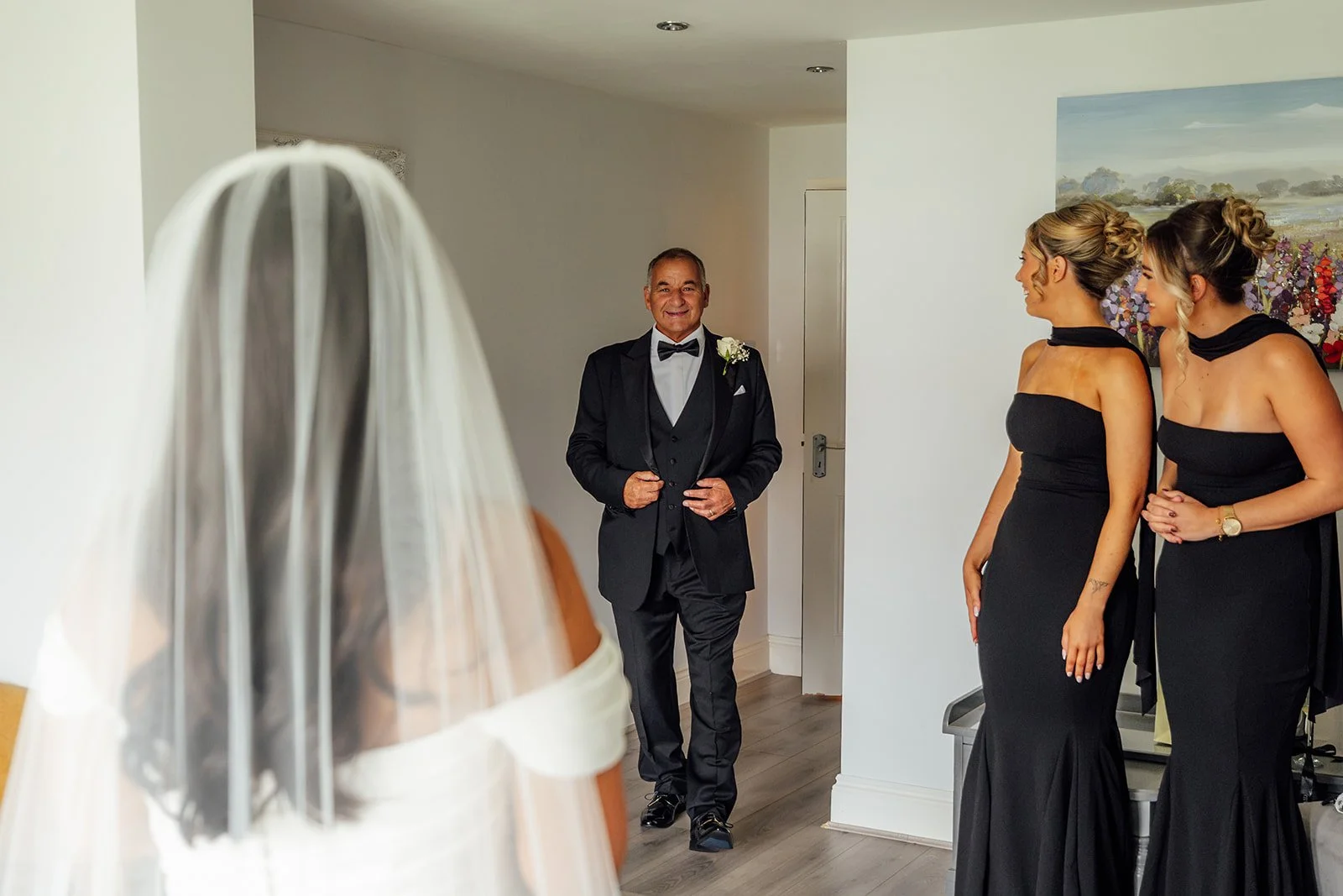 Nisha’s father smiles as he sees her in her wedding dress and veil, with bridesmaids in black dresses watching beside him during preparations for Nisha and Jack’s wedding at Batch Country House in Somerset, UK, photographed by Just James Weddings.