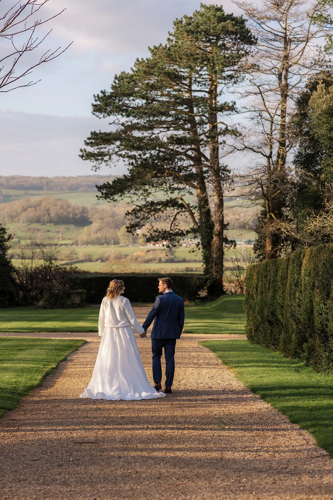 Jessie and Thomas walk hand in hand along the tree-lined grounds of Coombe Lodge in Somerset, UK, with sweeping Somerset countryside views behind them.