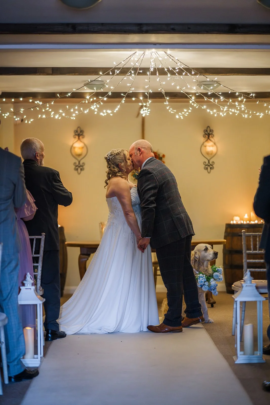 Debbie and Nigel share their first kiss during their indoor wedding ceremony at Yarlington Barn in Somerset, with fairy lights above, guests watching, and their golden retriever beside them in a blue floral collar.