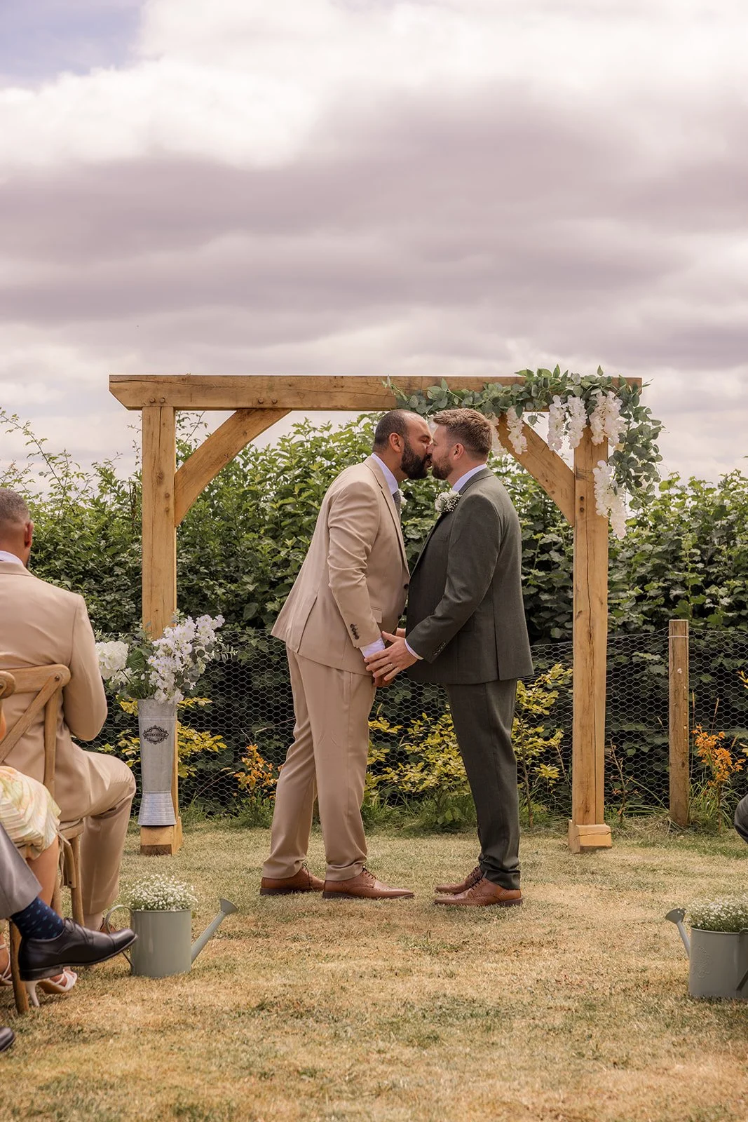 Aaron & Jack share their first kiss as a married couple beneath the flower-covered wooden arch during their outdoor ceremony at Rock Village Hall in Worcestershire, photographed by Just James Weddings.