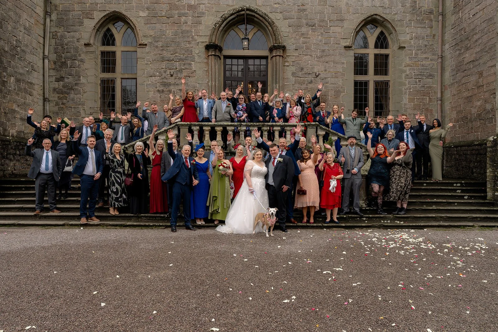 Becky & Rob stand with family and guests on the grand steps of Clearwell Castle in Gloucestershire, UK for a large wedding group photo. The couple and their dog are surrounded by smiling guests outside the historic Gloucestershire wedding venue.
