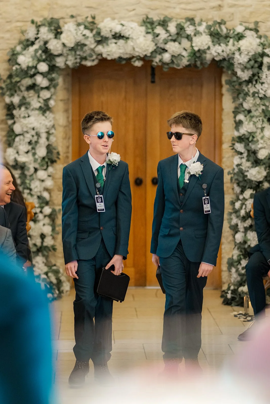 Two young ushers in blue suits and sunglasses walk down the aisle beneath a white floral arch at Kingscote Barn in the Cotswolds, UK, during Siobhan & Nicholas' wedding ceremony, a fun rustic barn wedding moment captured before the couple enter.